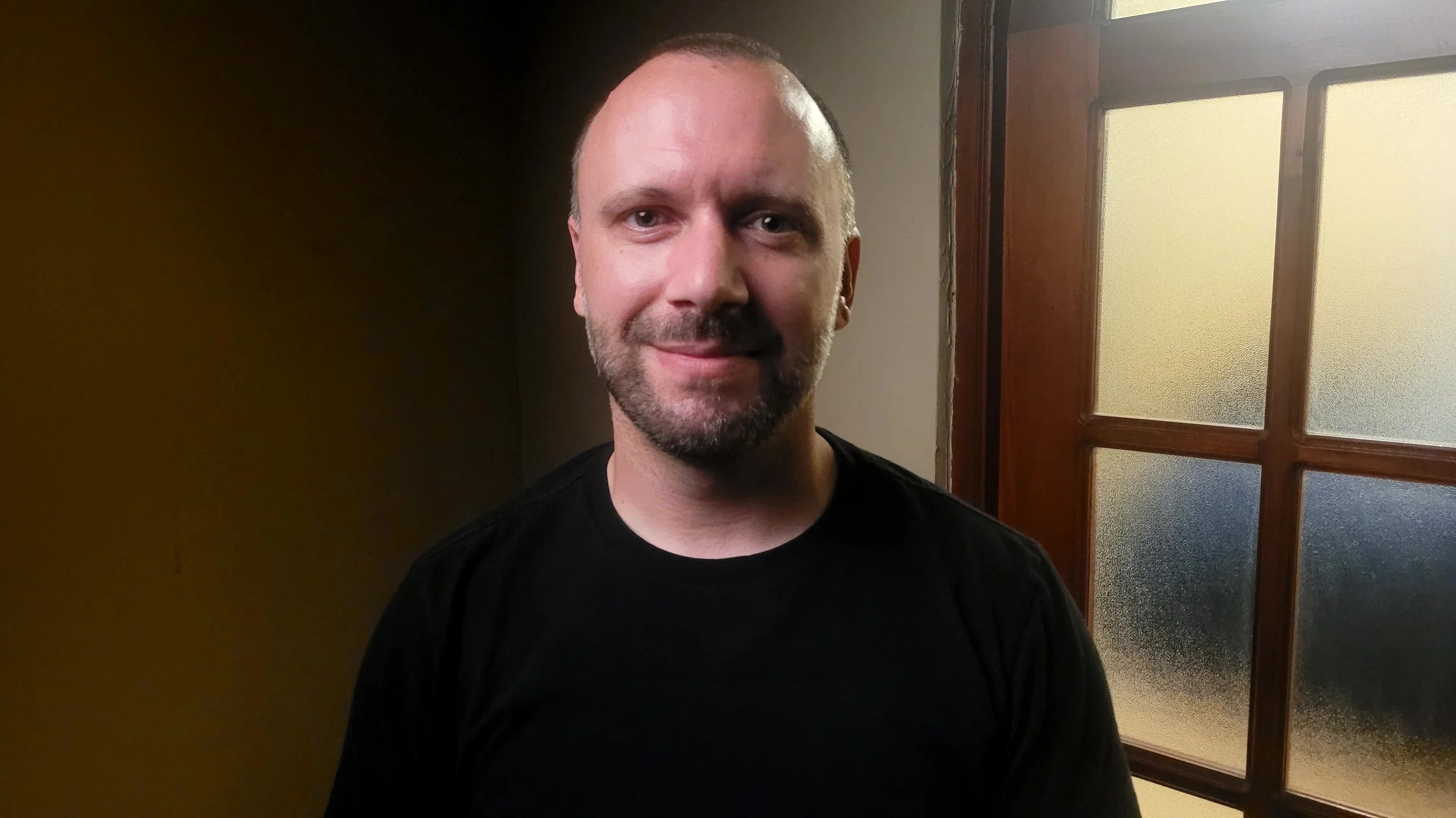 Darren Soares, smiling with a beard and short hair, wearing a black shirt, standing in front of a door with glass panes. DarrenSoares.com - A Humans Guide To Healing
