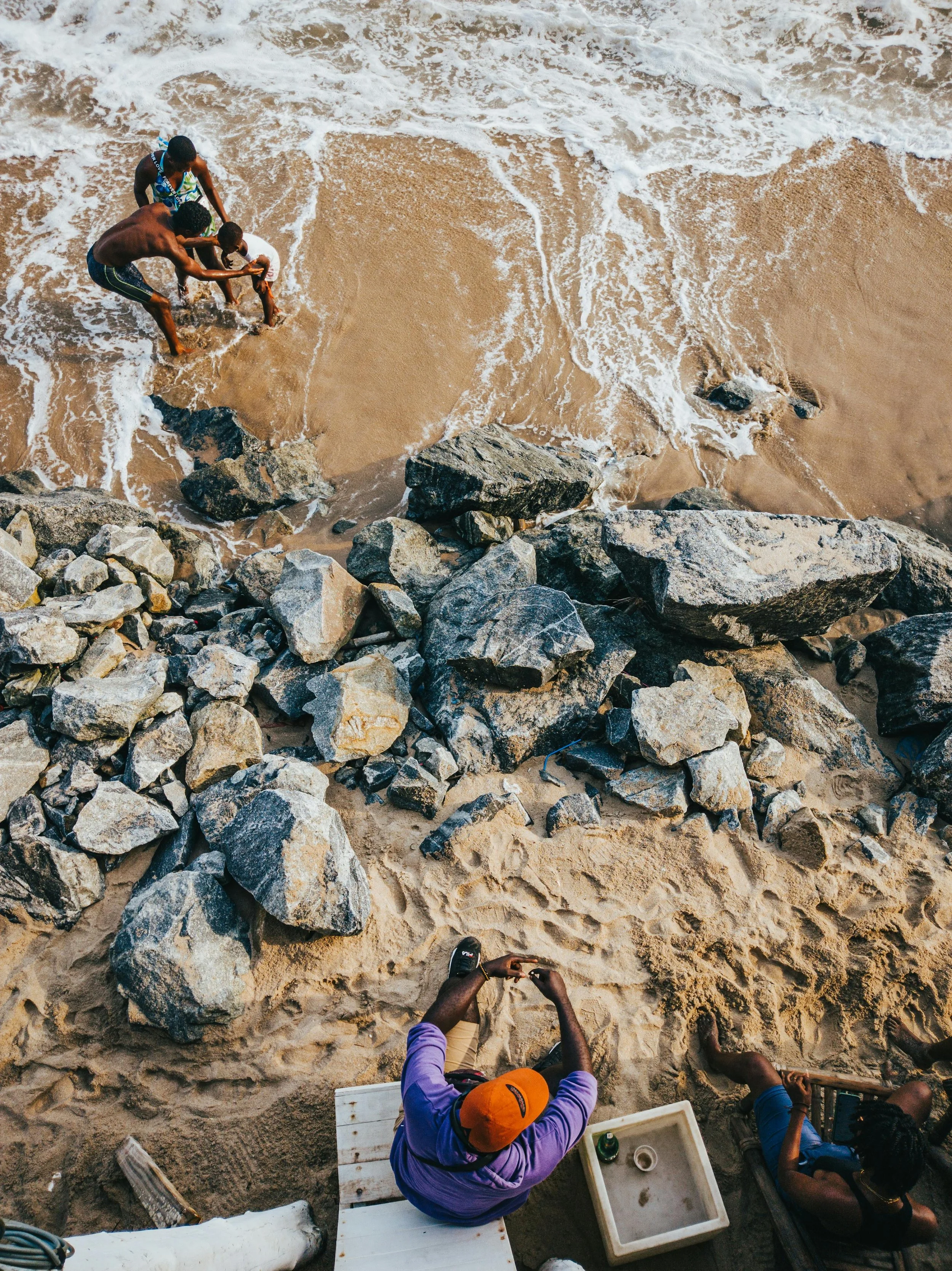 People on a rocky beach with rocks, sand, and ocean waves, some are playing in the water, and two individuals are sitting under a shaded area.