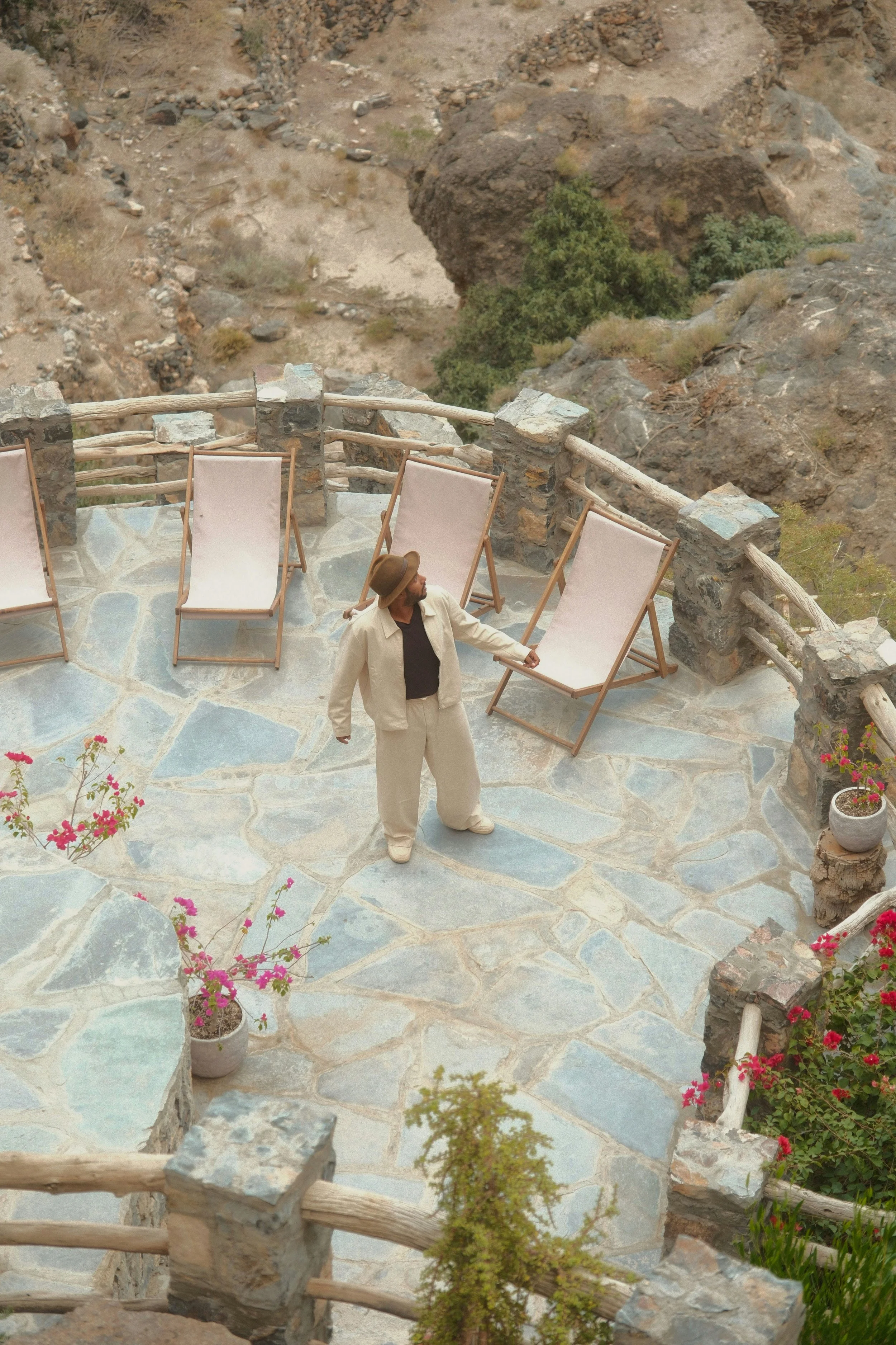 A man dressed in beige clothes and a hat is standing on a stone patio with several pink deck chairs, overlooking a rugged, rocky landscape with sparse vegetation and flowering plants in pots.