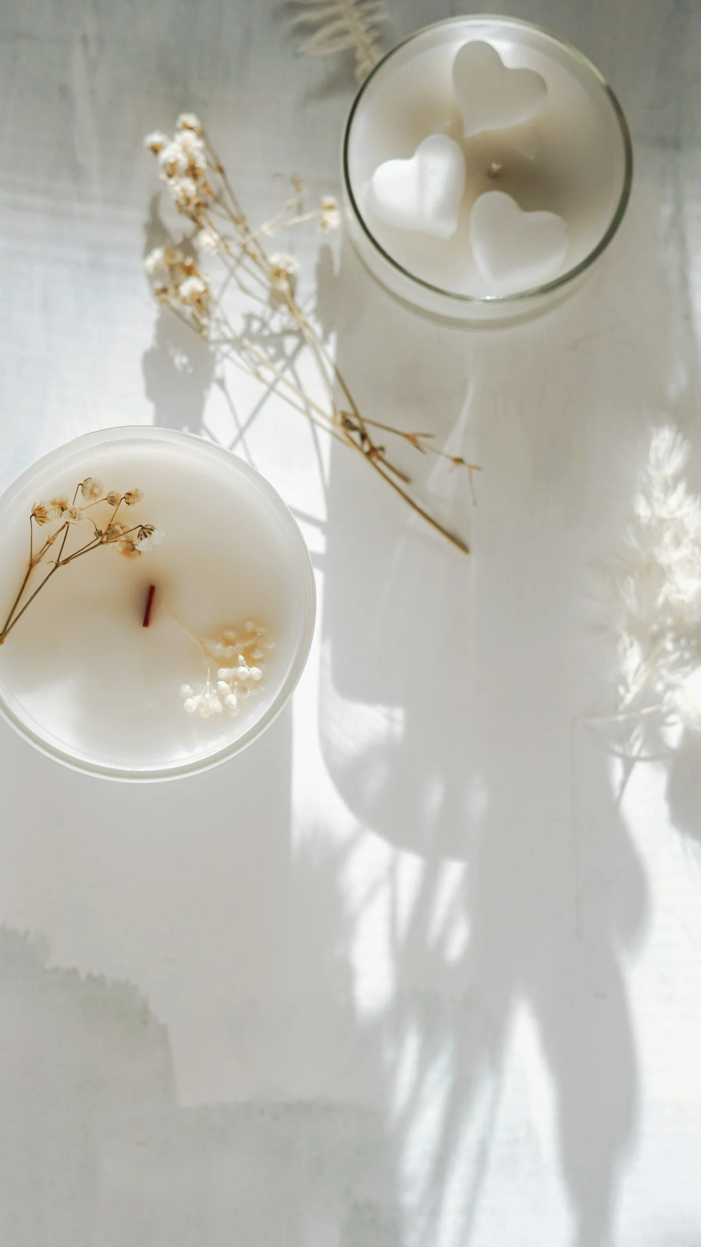Two white candles with dried flowers inside and three white heart-shaped wax melts floating in a glass. Dried flowers are also placed on a white surface, casting shadows.