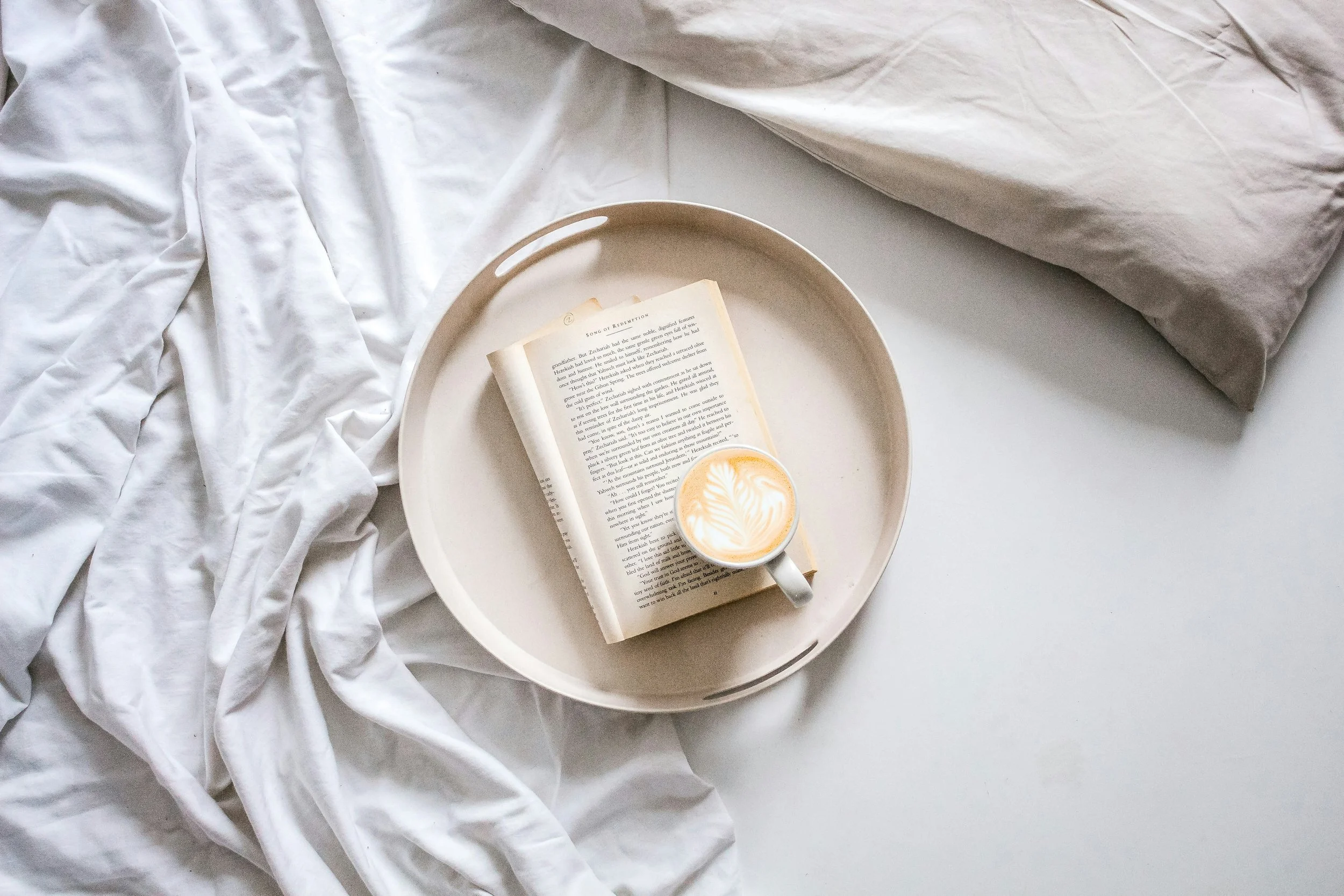 A book and a latte on a white tray on a bed with crumpled white sheets and a beige pillow.