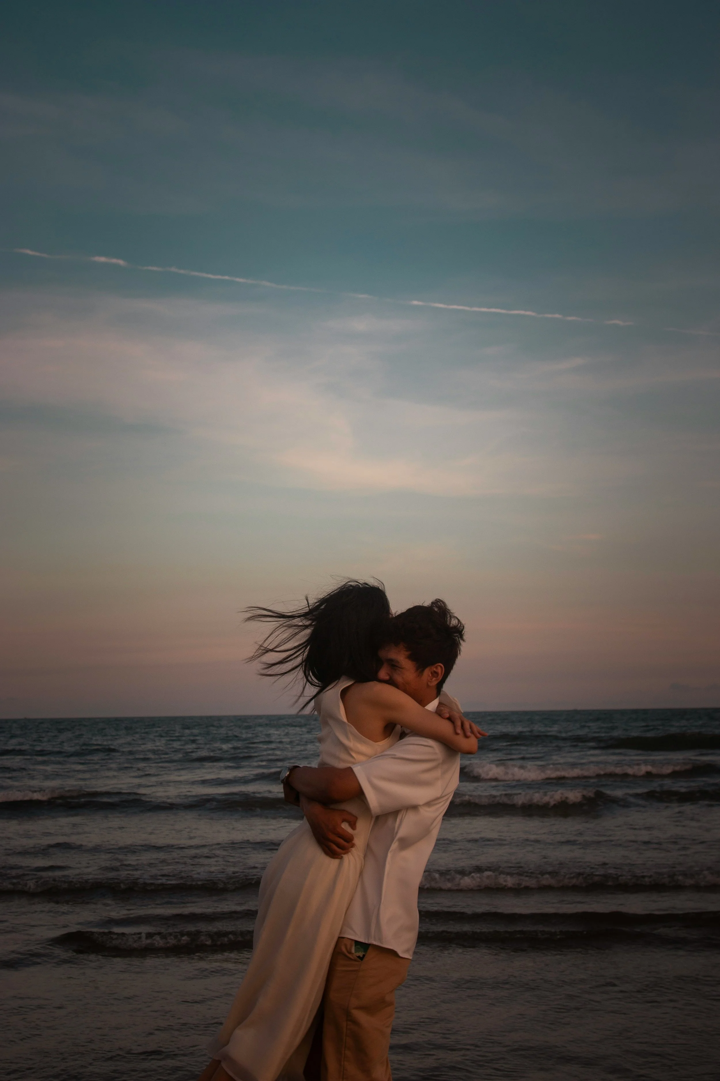 A man and woman embracing on a beach at sunset, with the ocean and sky in the background.