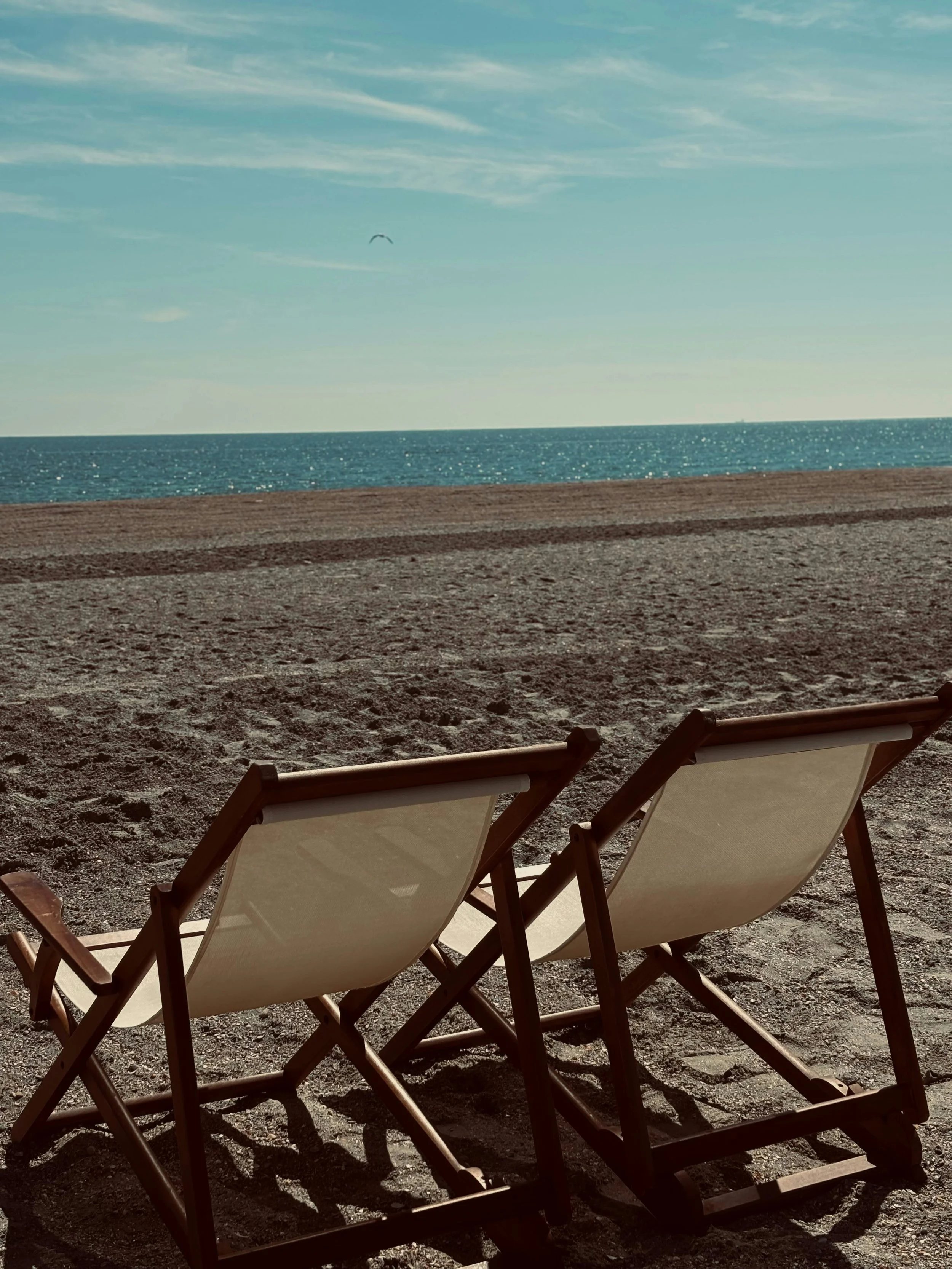 Two empty lounge chairs on a sandy beach facing the ocean with a clear blue sky and a single kite in the distance.
