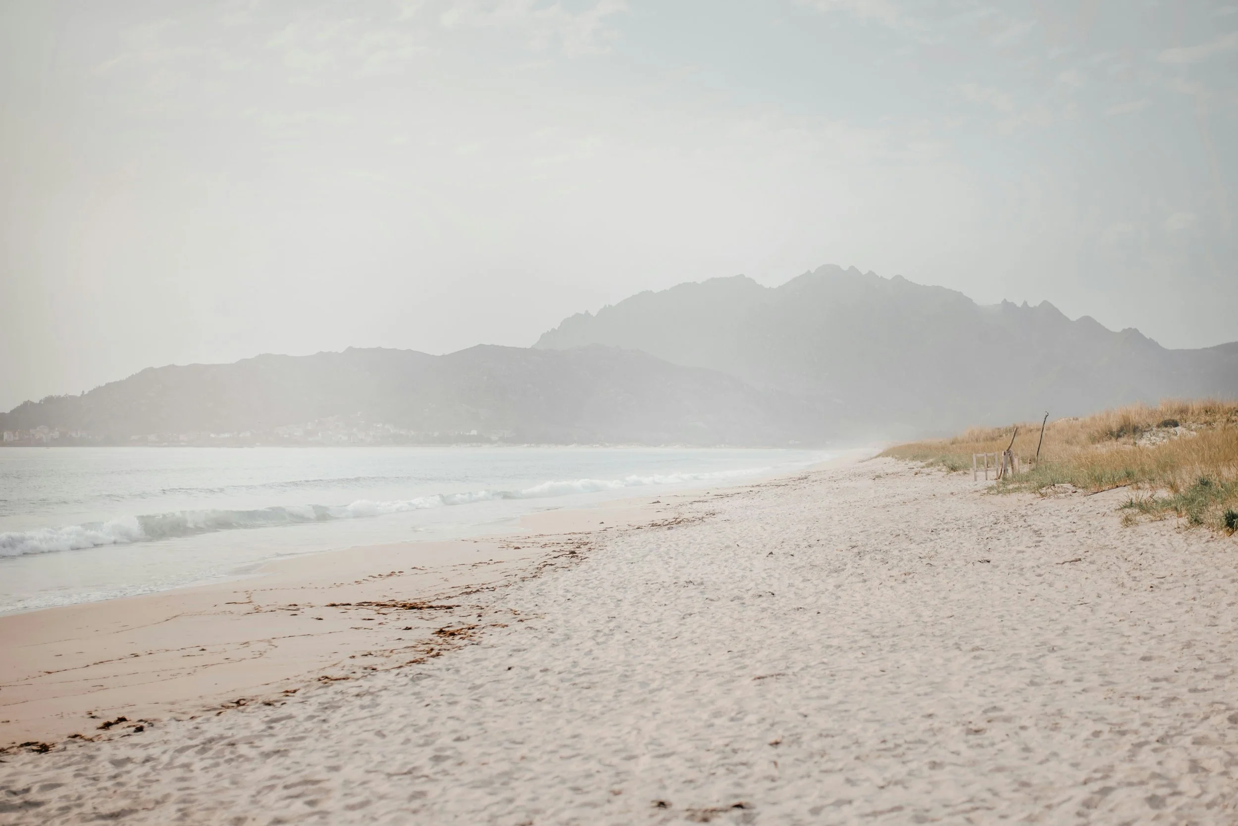 Empty sandy beach with gentle waves and distant mountains under an overcast sky.