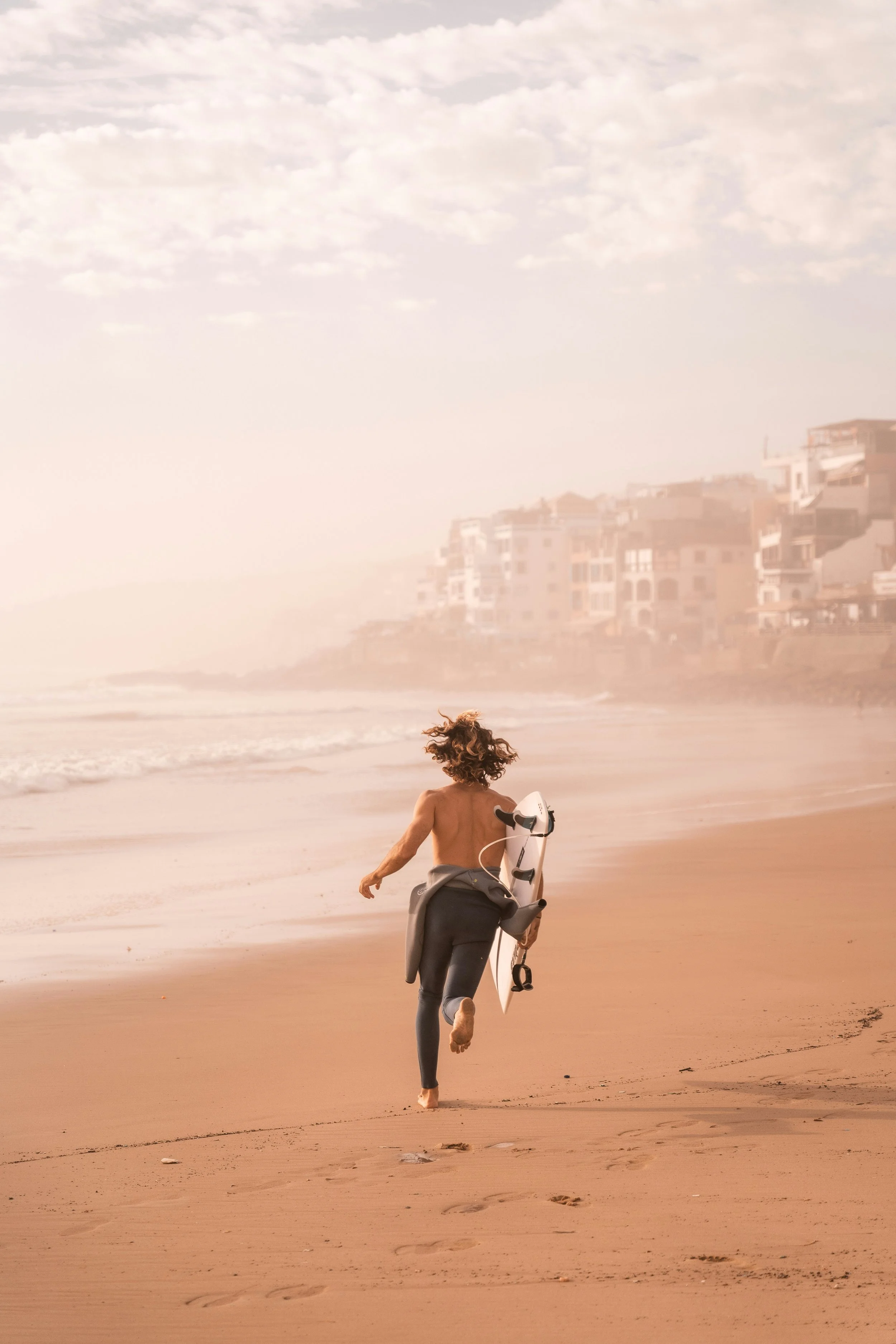 A woman running on a sandy beach carrying a surfboard, with a foggy shoreline and buildings in the background.