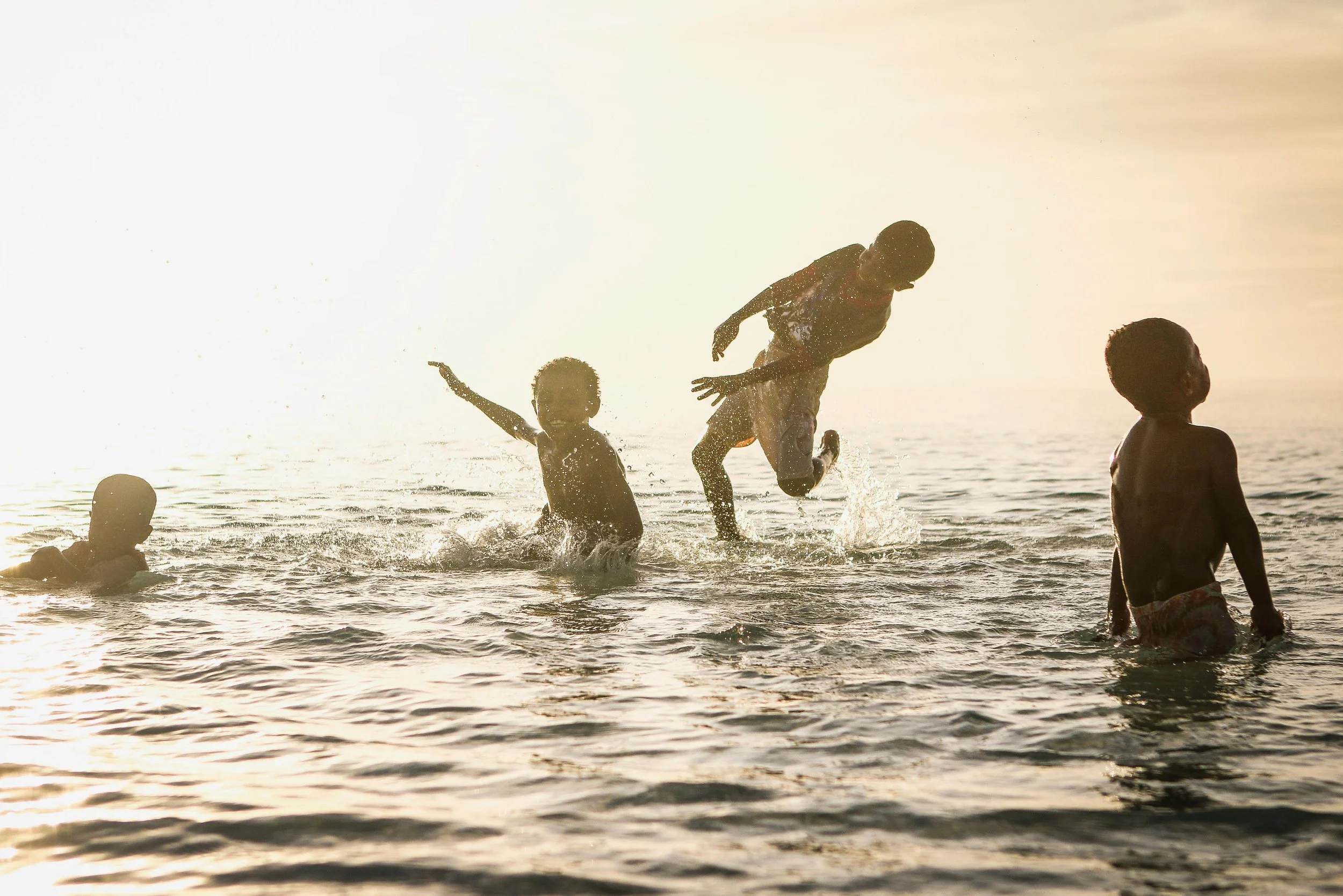 Four children playing and splashing in the water at sunset or sunrise, with the sky and horizon in the background.
