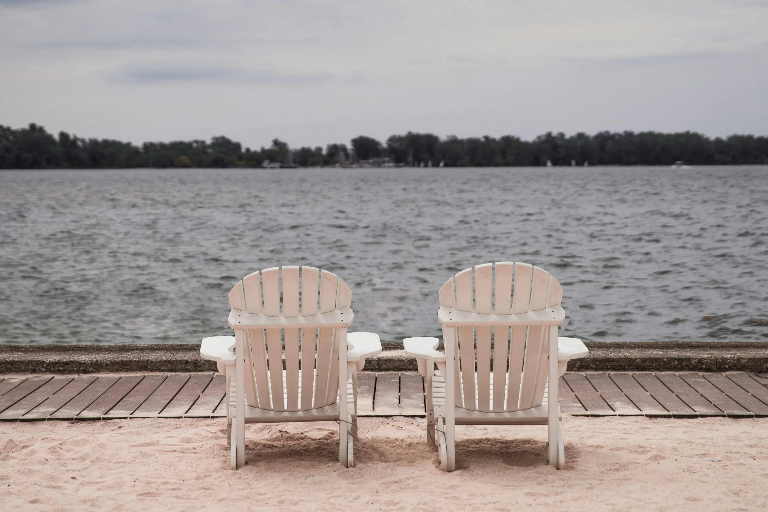 Two white Adirondack chairs facing a lake, with a wooden boardwalk and sandy beach in the foreground and trees on the distant shoreline under a cloudy sky.