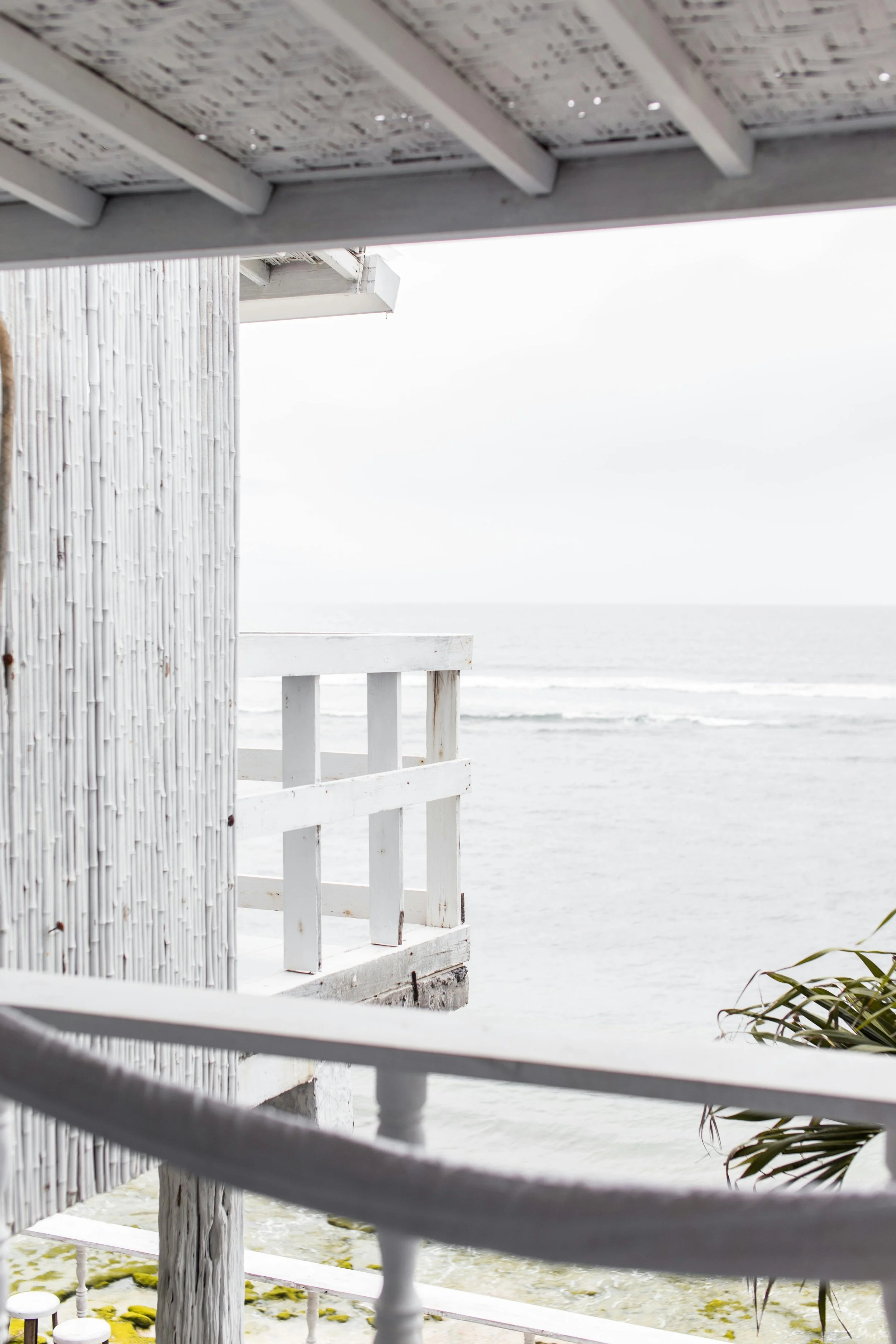 View from a beach house balcony showing white wooden railing, part of the exterior wall, and the ocean in the background on a cloudy day.