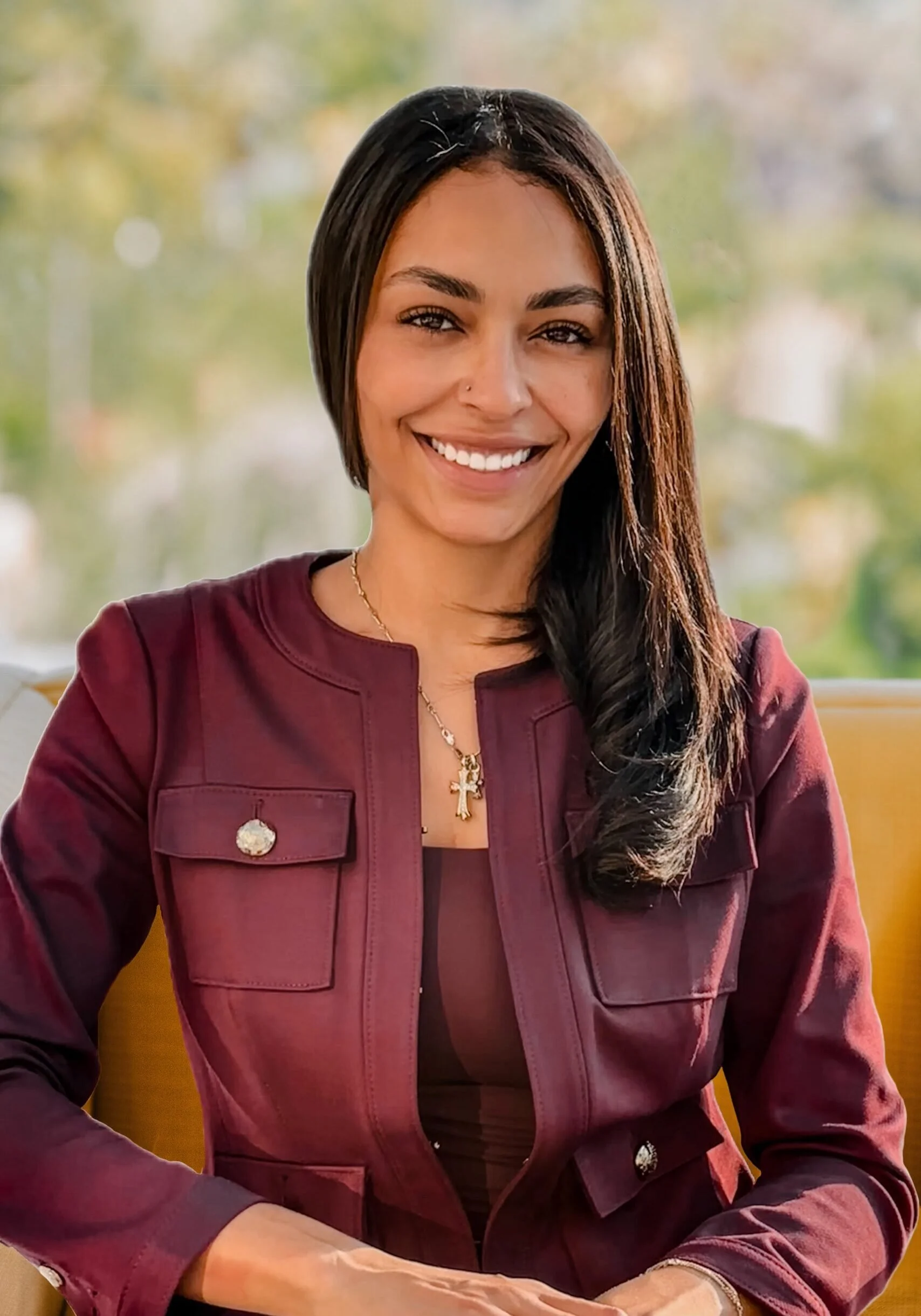 A woman with long dark hair, wearing a maroon jacket and a cross necklace, sitting indoors in front of a window with blurred trees outside, smiling at the camera.