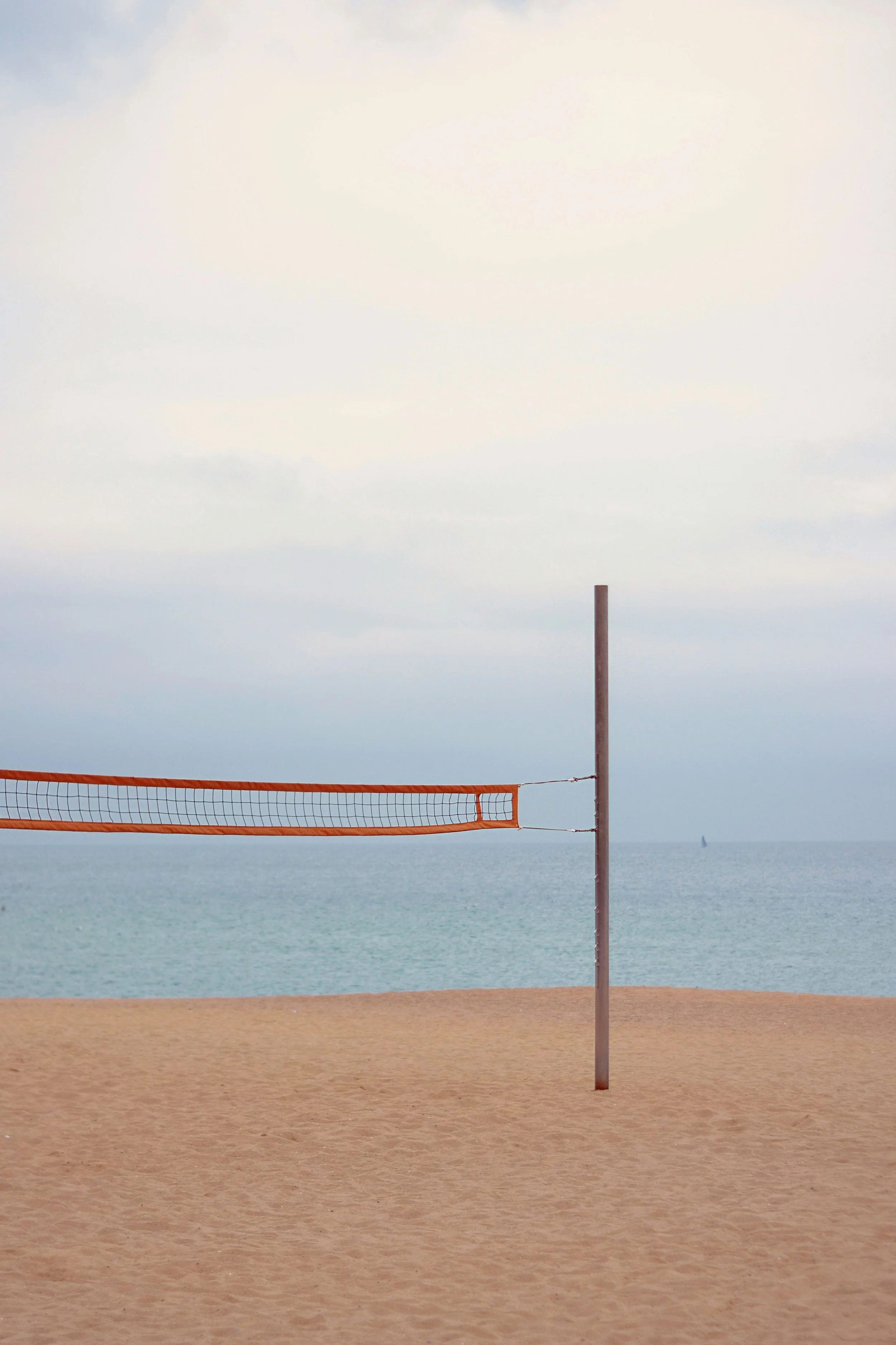 Empty beach volleyball court with a net set up, sandy ground, ocean in the background under a cloudy sky.