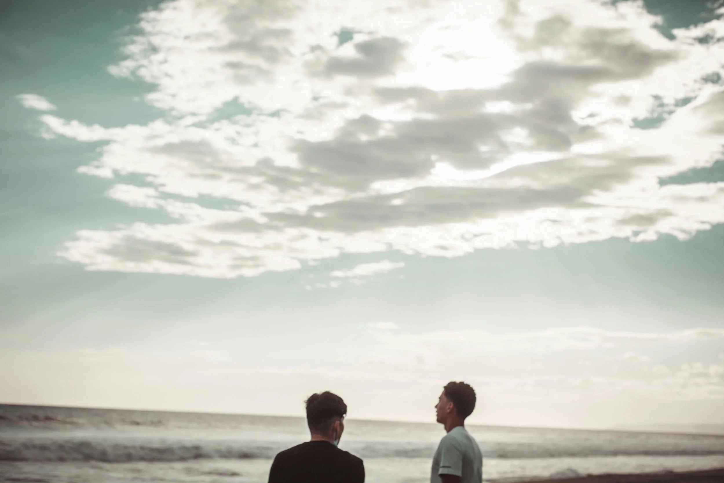 Two men standing on the beach looking at the ocean under a cloudy sky during sunset or sunrise.
