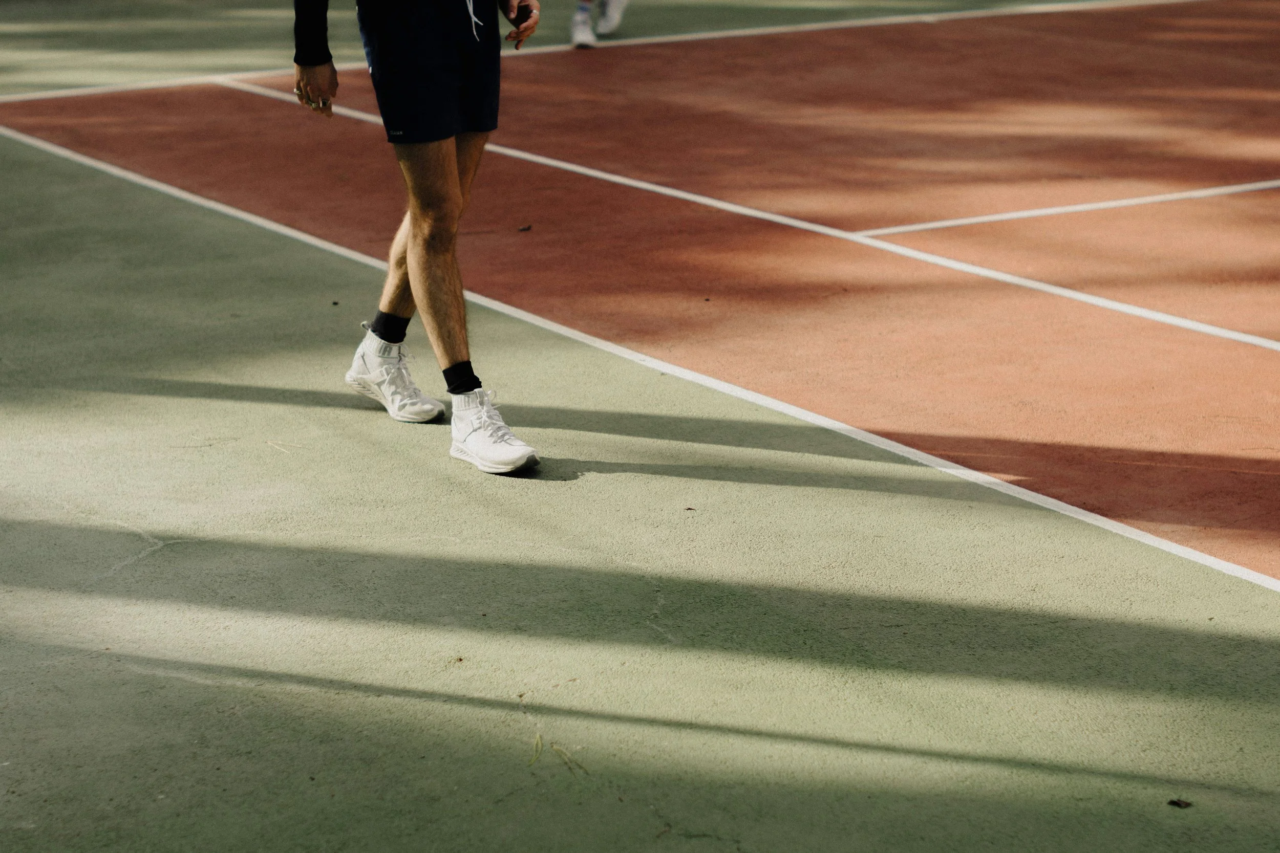 Partial view of a man walking on a tennis court, wearing white sneakers, black shorts, and a black top. The court has green and red sections with white lines.