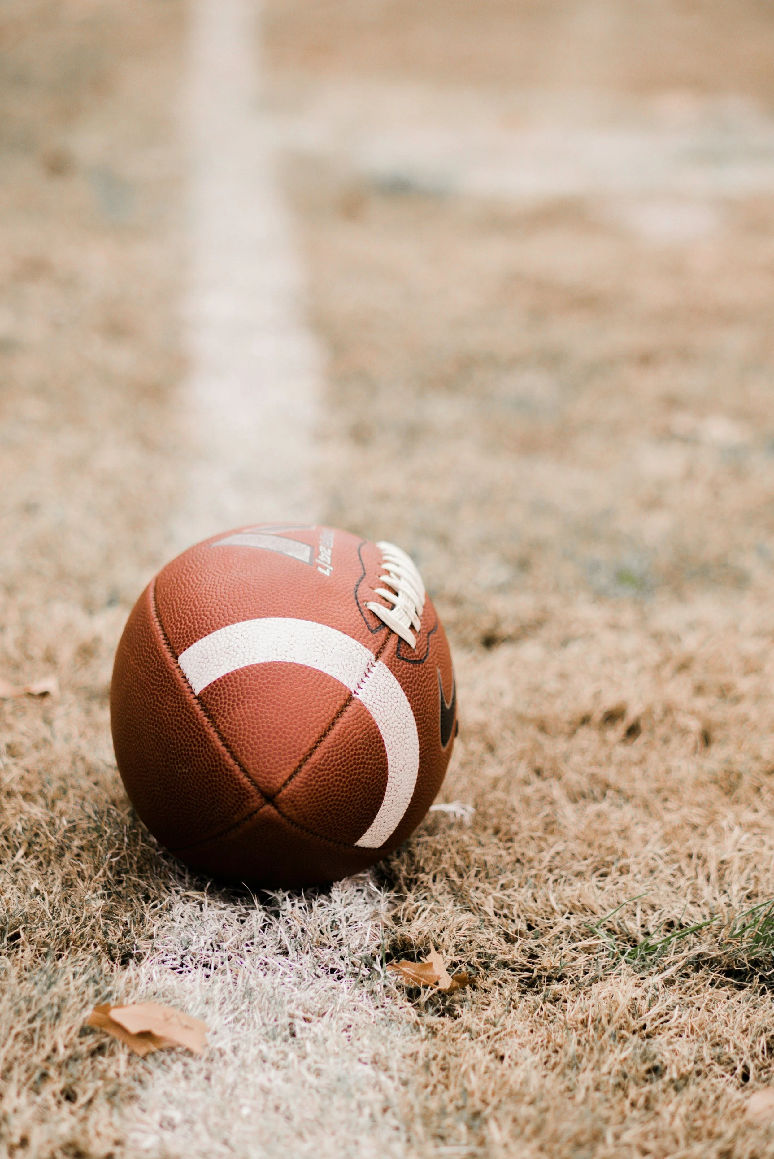 An American football resting on a dry, grassy field with white yard line markings.