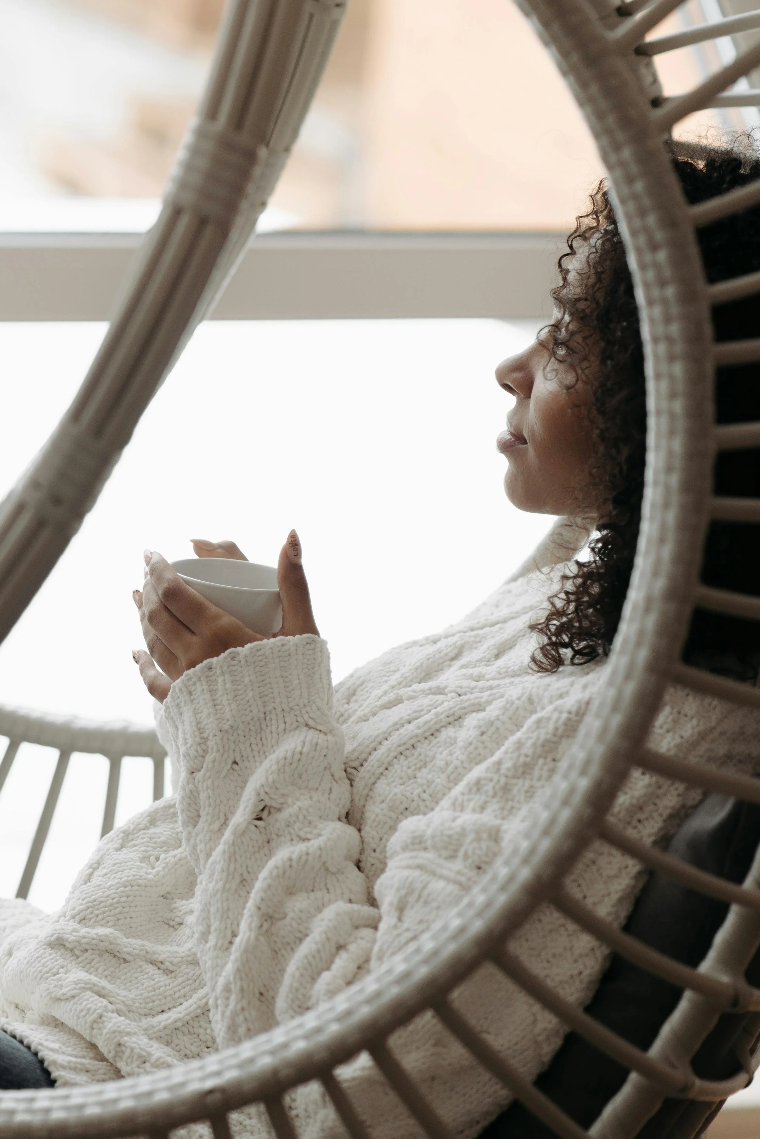 A woman with curly hair wearing a cream-colored knitted sweater, sitting in a circular wicker chair, holding a white mug, looking thoughtfully out a window.