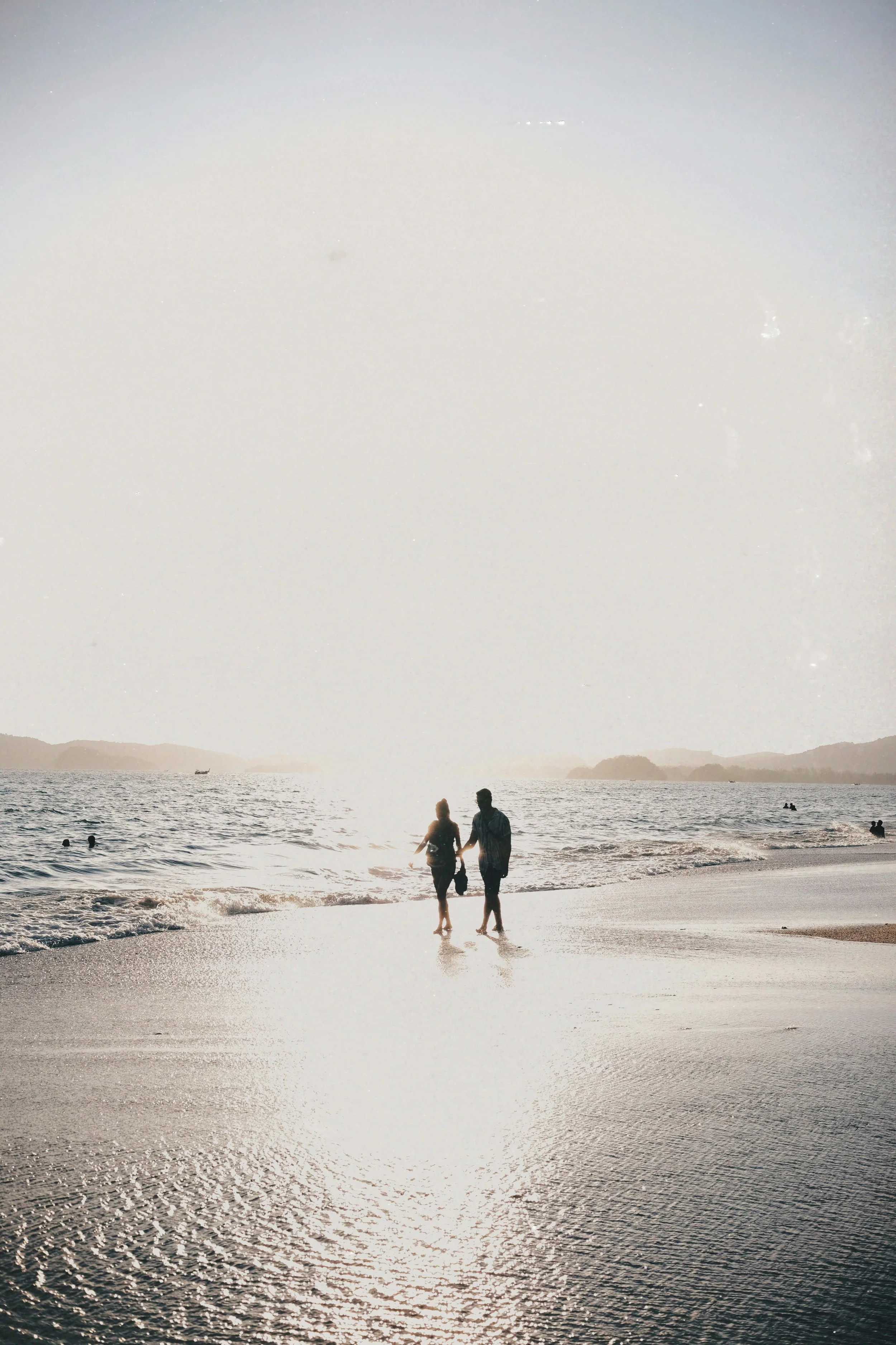 Silhouettes of a couple walking on the beach holding hands during sunset or sunrise, with ocean waves and distant islands or landforms in the background.