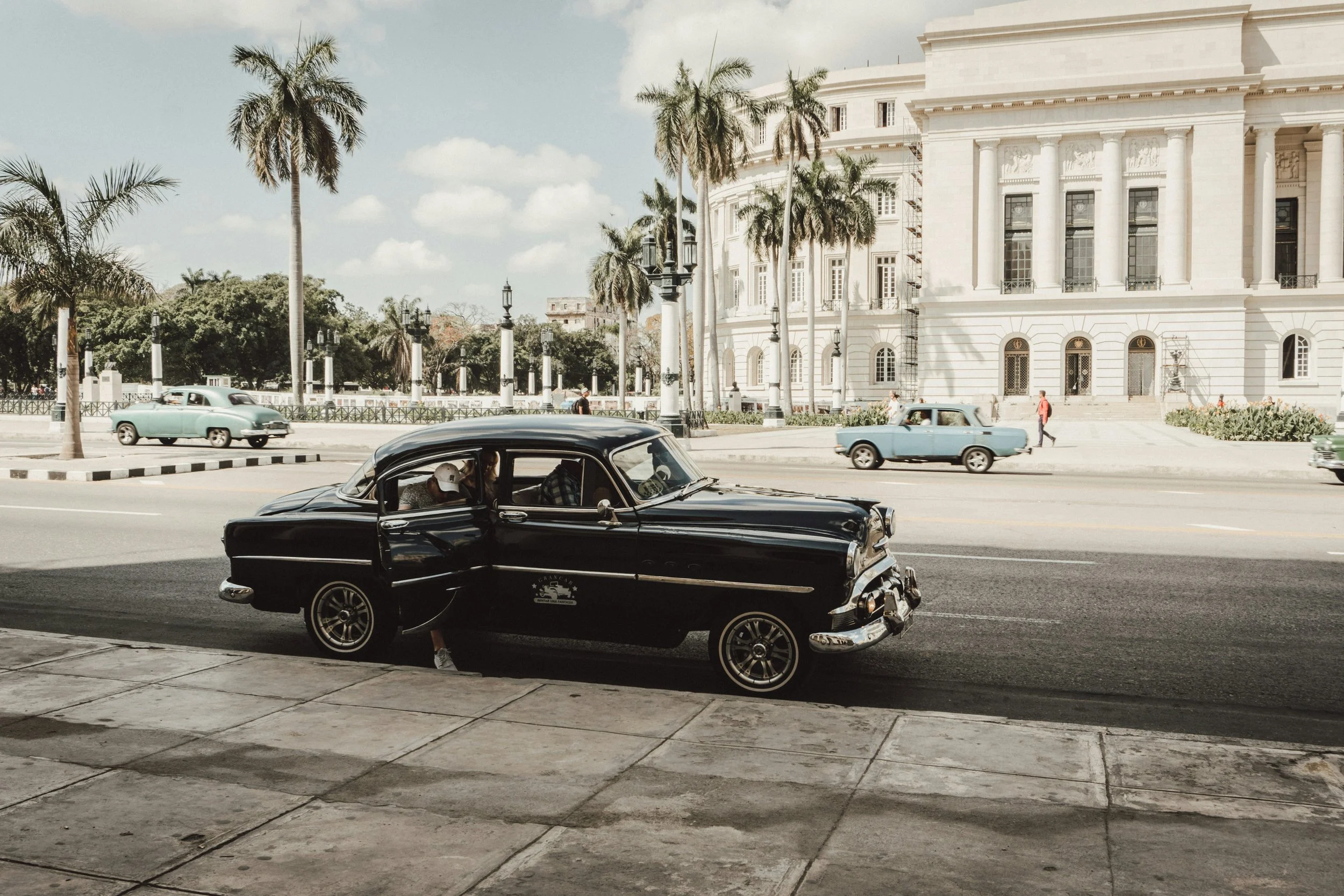 A black vintage car parked on the side of a street with its door open, in front of a historic white building with columns, surrounded by palm trees, with a few other vintage cars driving and pedestrians walking in the background.
