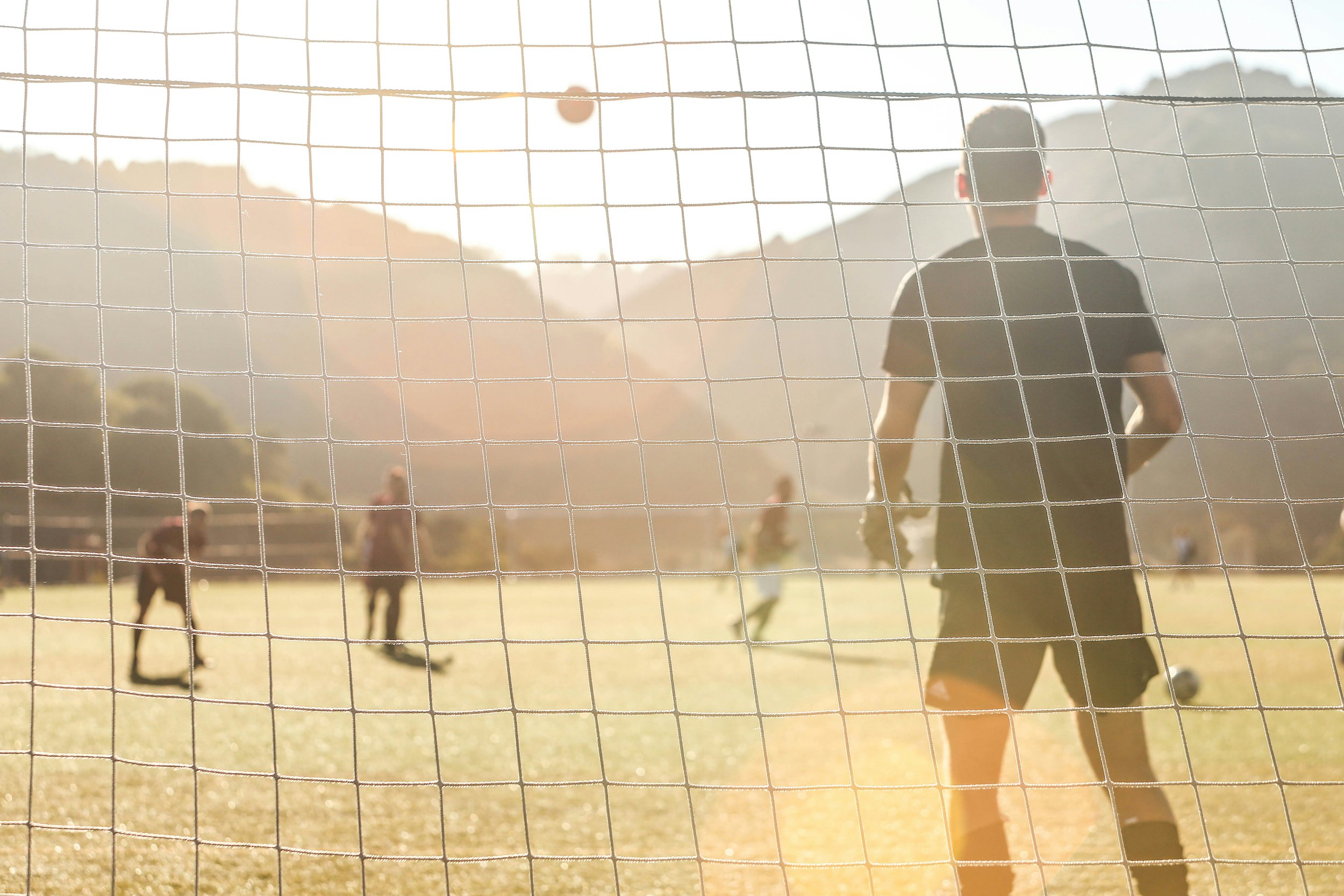 Soccer players practicing on a field during sunset, viewed through the goal net.