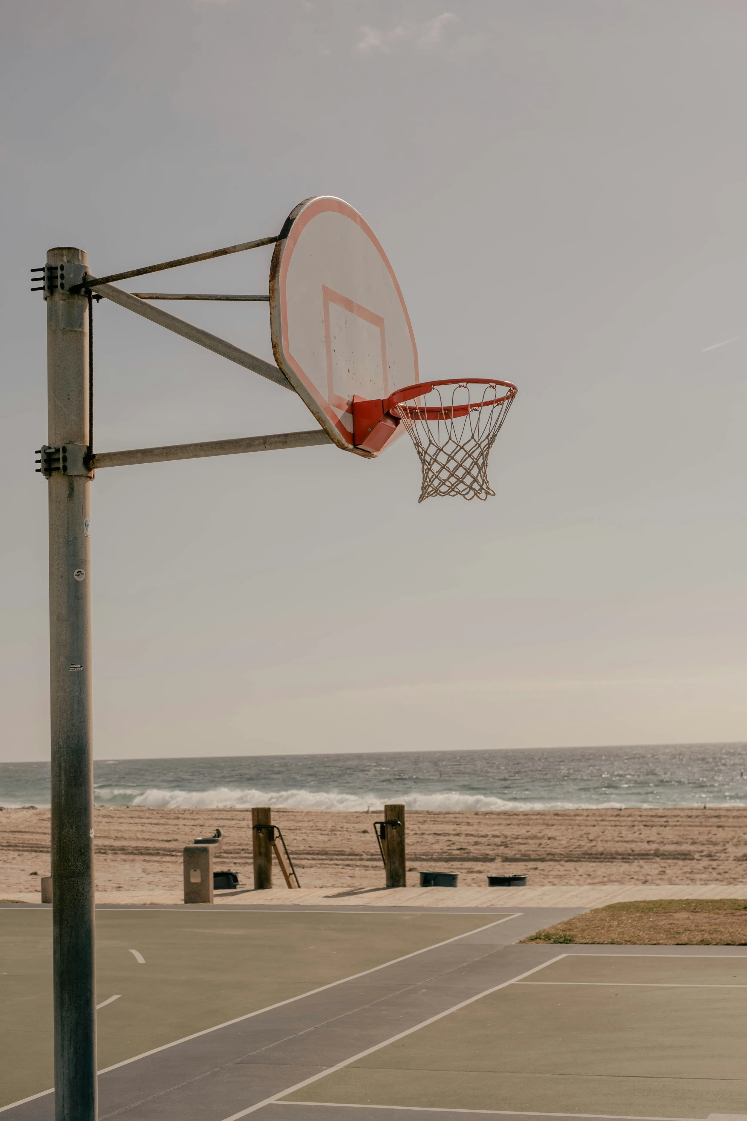 Empty outdoor basketball court with a basketball hoop, set against a beach and ocean background, under a cloudy sky.