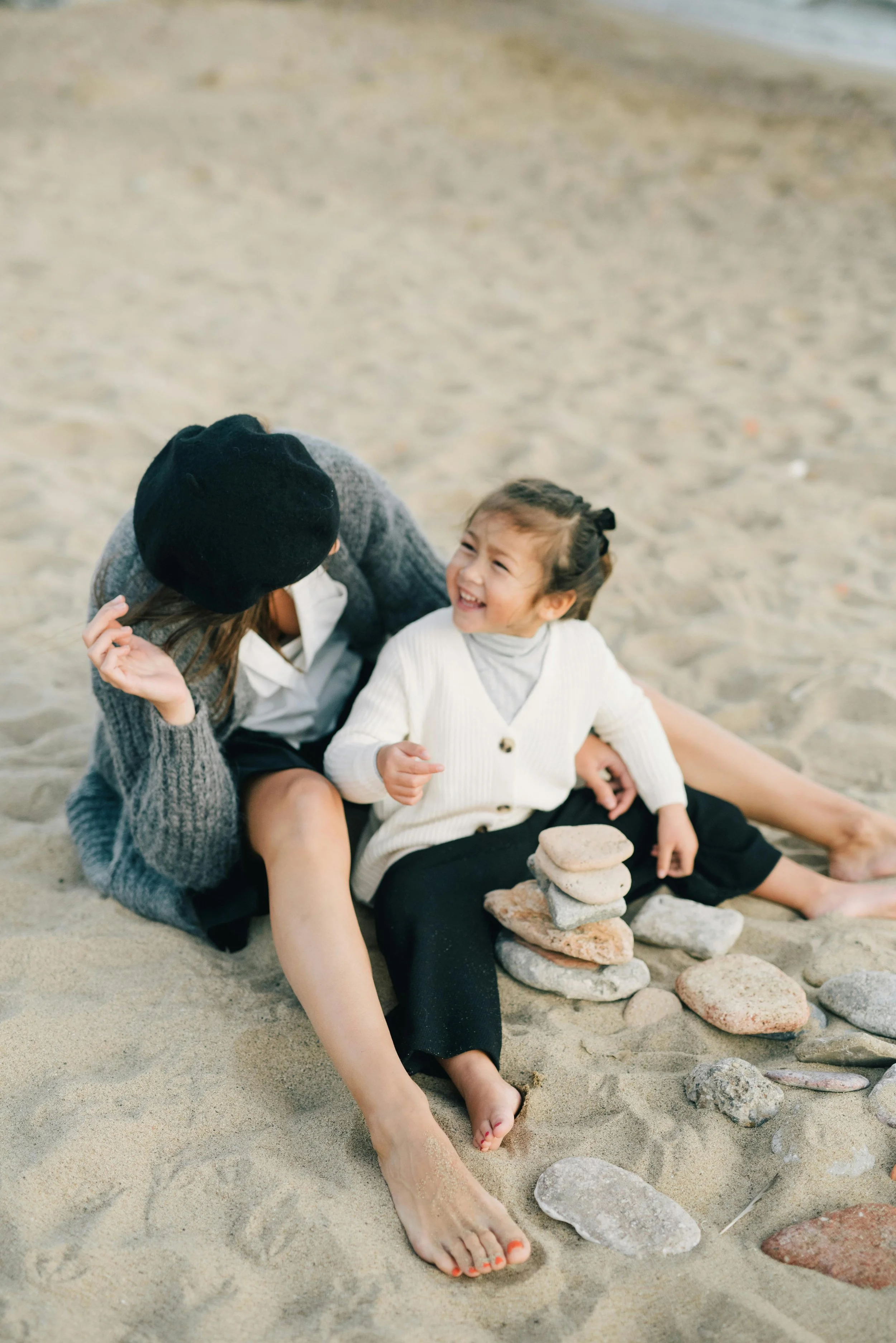 A woman and a young girl sitting on the sand at the beach, smiling and laughing together. The woman is wearing a black hat and a gray cardigan, while the girl is in a white sweater and black pants, with a stack of stones in front of her.