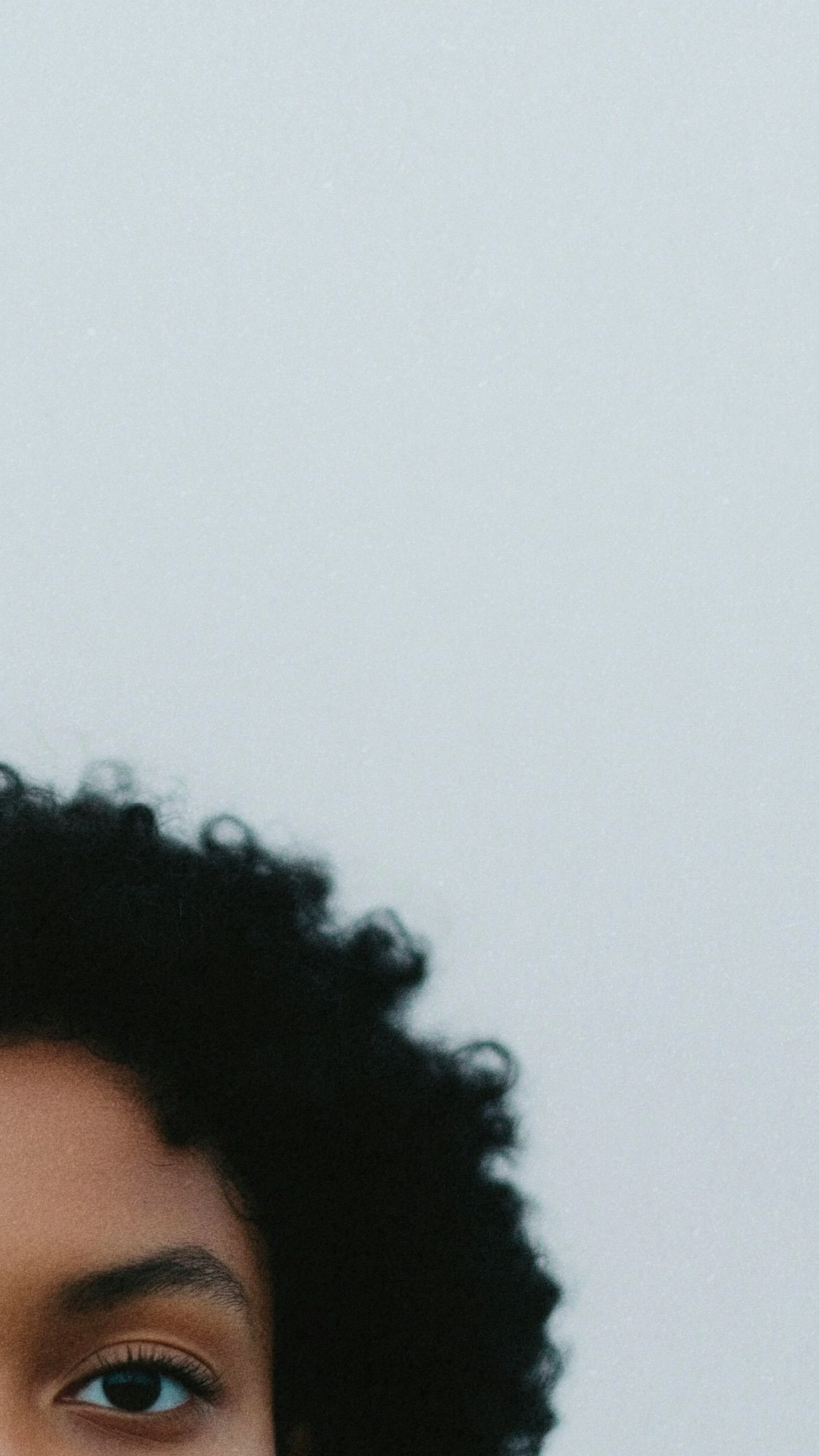 Close-up of a person with natural curly hair and brown eyes, capturing part of their face and hair against a plain gray background.