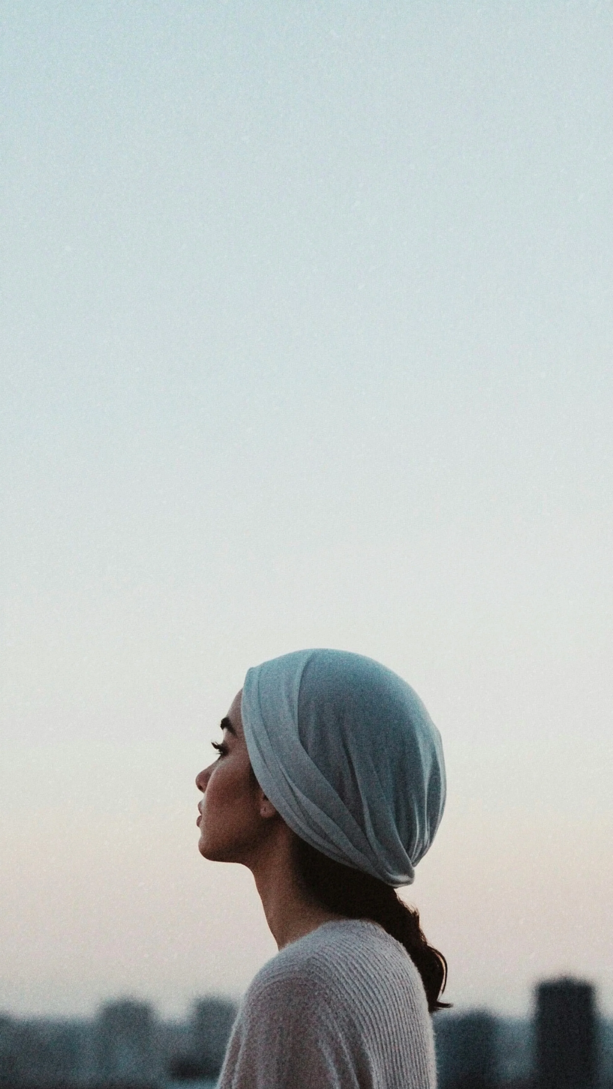 Profile of a woman with a headscarf, standing outdoors against a dusk sky with city buildings in the background.