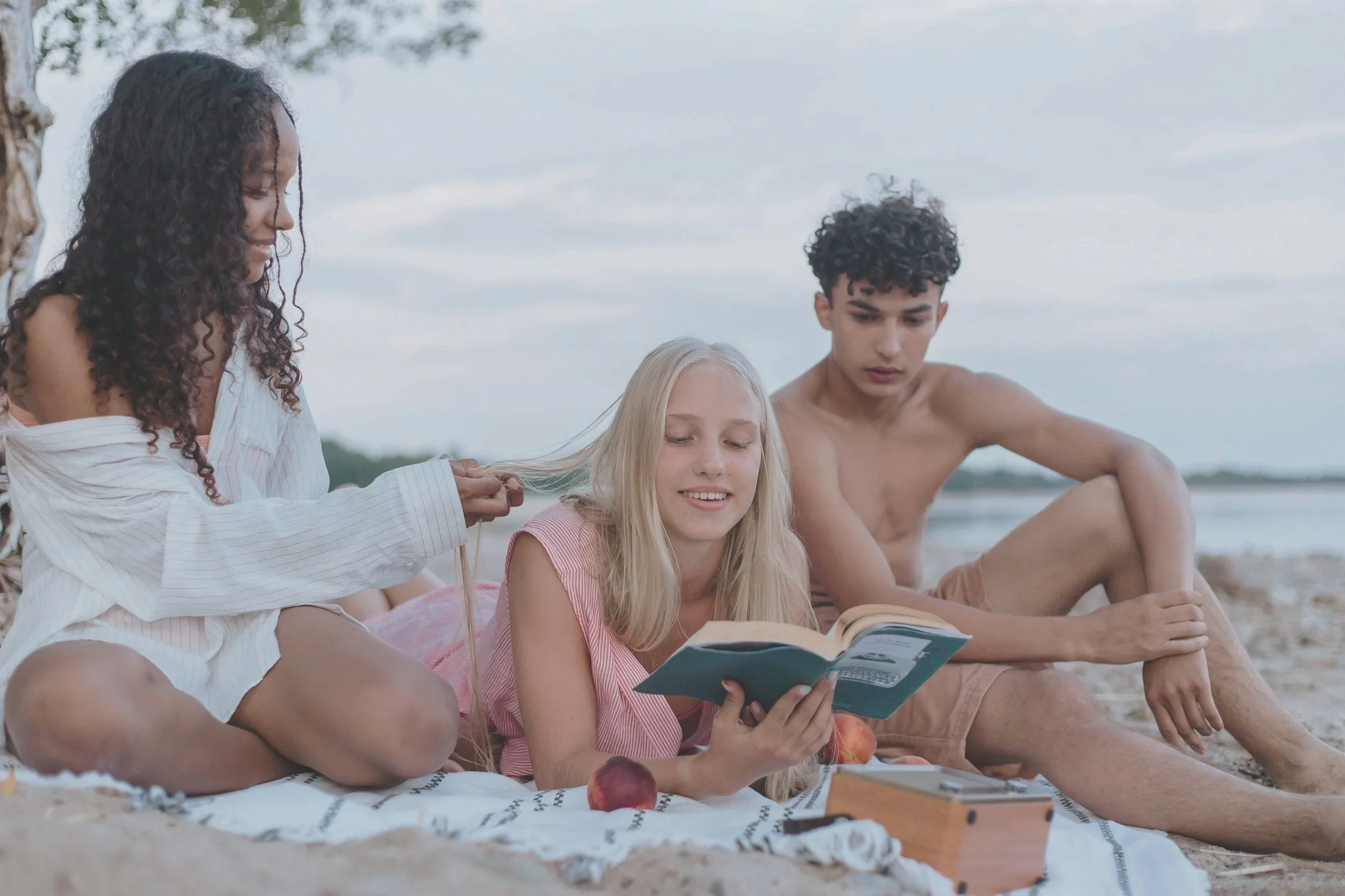 Three young people sitting on a beach blanket, with one girl reading a book and another girl holding a braid, while a shirtless boy looks at the book, with sand and water in the background.