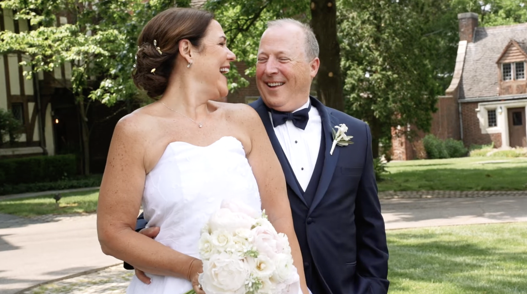 A bride and groom smiling and enjoying their wedding day outdoors, with trees and a house in the background.