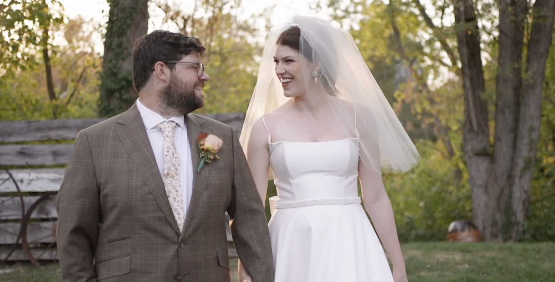 A bride and groom holding hands, smiling at each other outdoors during a wedding ceremony, with trees and autumn foliage in the background.