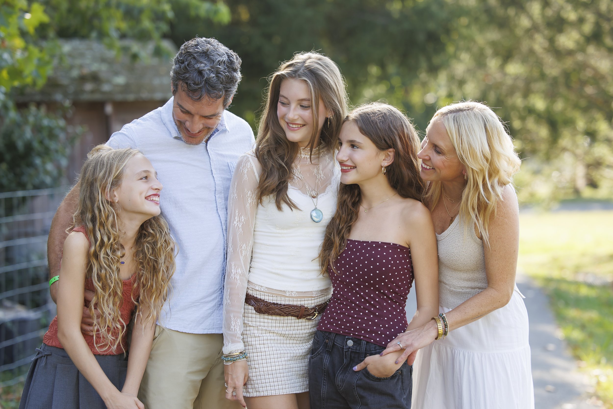 A family of six smiling and interacting outdoors on a sunny day, standing on a pathway with green trees in the background.