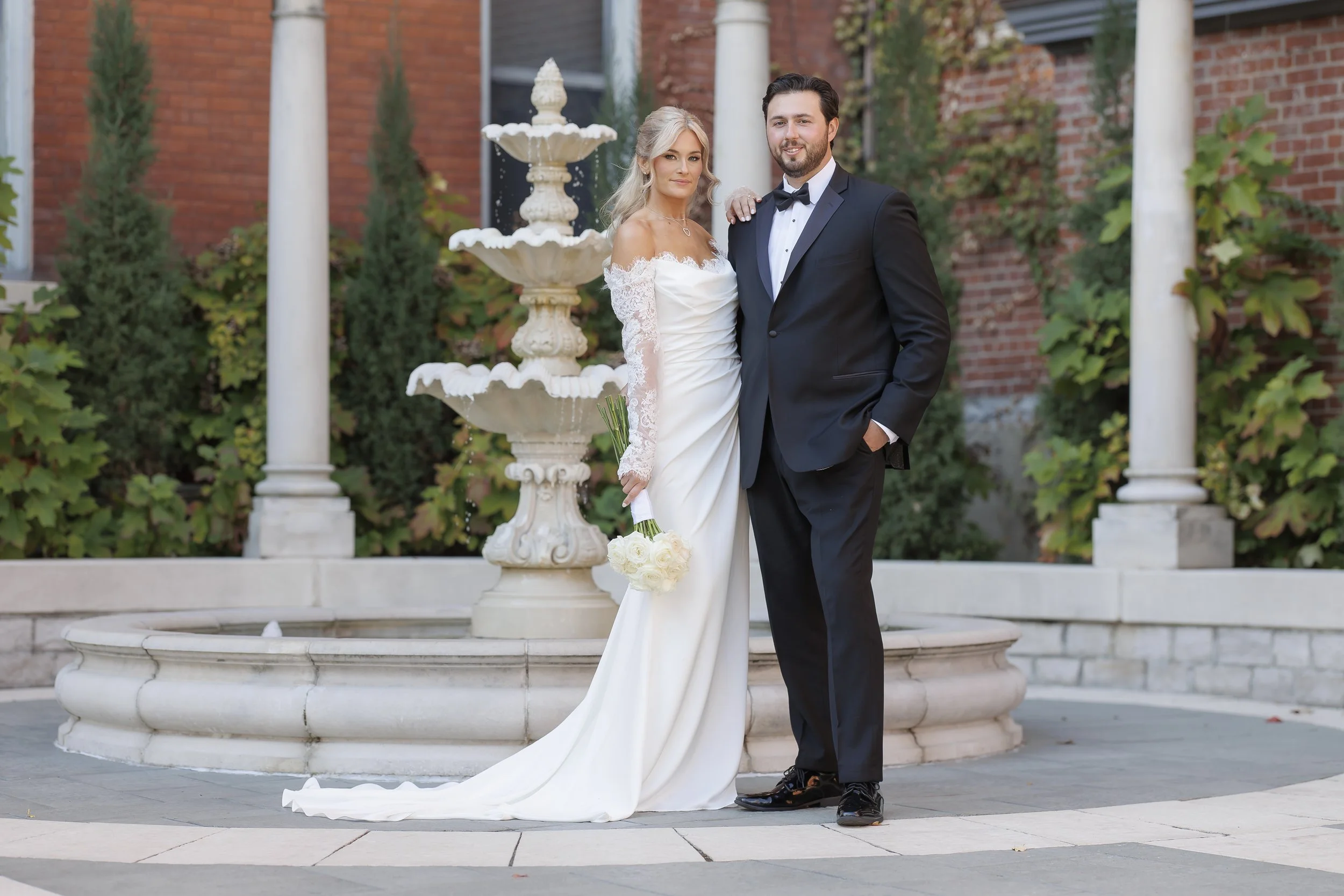 A bride and groom standing together outside near a fountain, with the bride holding a bouquet of white roses. The bride is in a white wedding dress and the groom is in a black tuxedo. Background features a brick building and greenery.