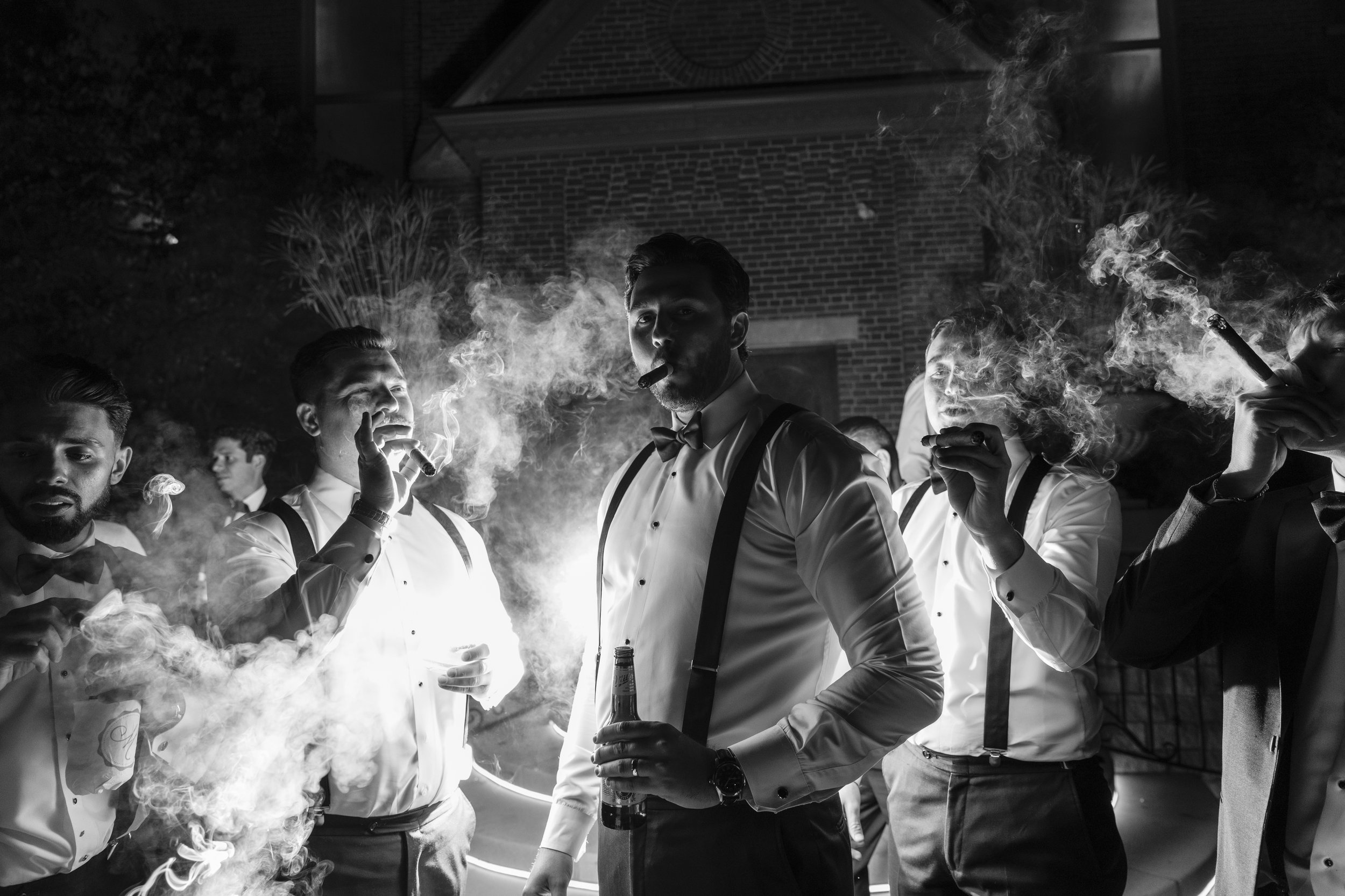 A group of well-dressed men in tuxedos, some smoking cigars, at night outside a building. One man in the center holds a bottle, and another is lighting a cigar. Bright light from a fire or spotlight illuminates the scene, casting shadows.