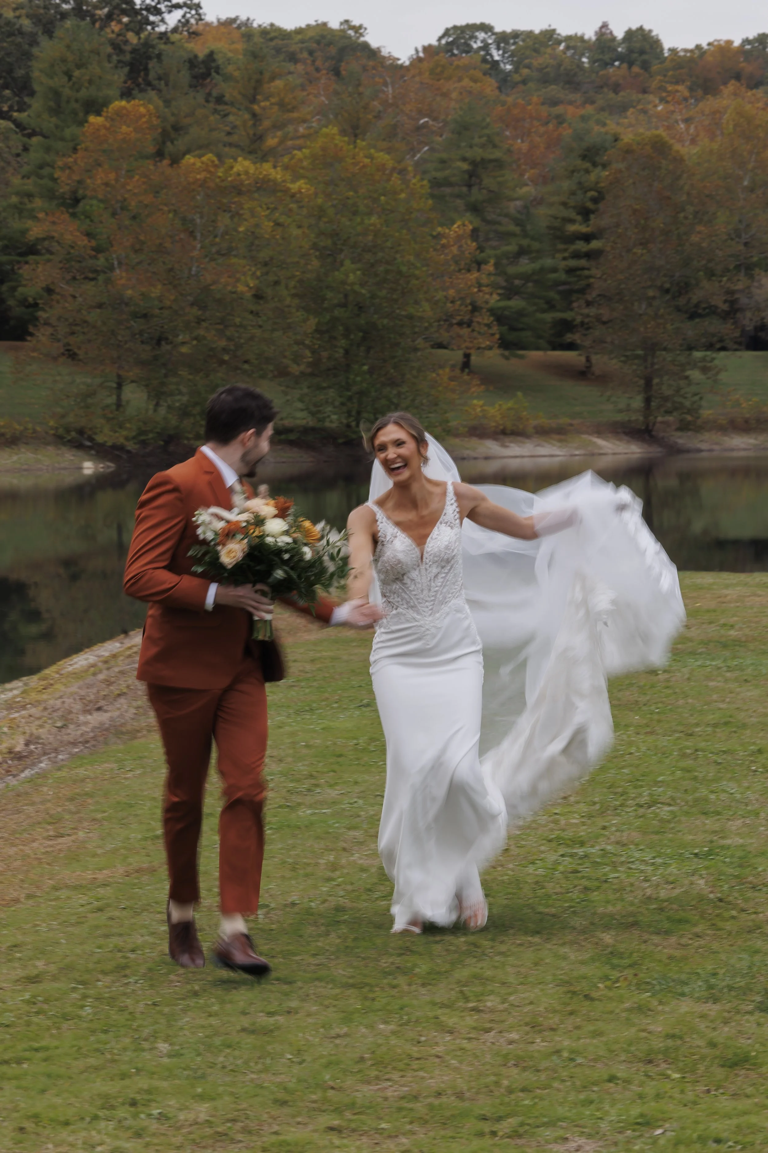 A joyful bride and groom running on a grassy area near a lake, with trees displaying fall foliage in the background. She is in a white wedding dress, and he is holding a bouquet of flowers.