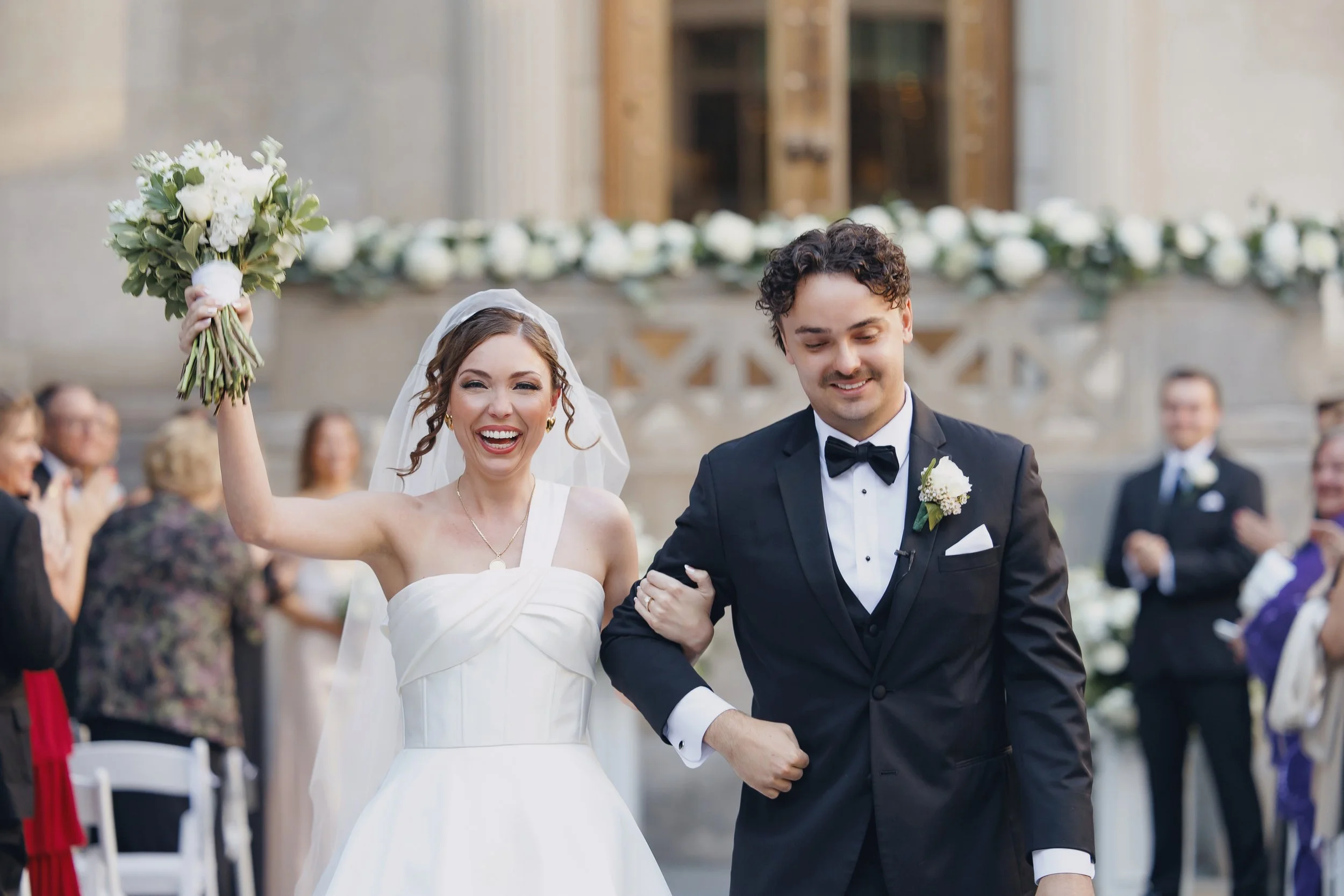 A bride and groom walking down the aisle at their wedding. The bride is smiling happily, holding a bouquet in her raised hand, and wearing a white wedding dress. The groom is smiling and dressed in a black tuxedo with a bow tie. Guests are clapping i