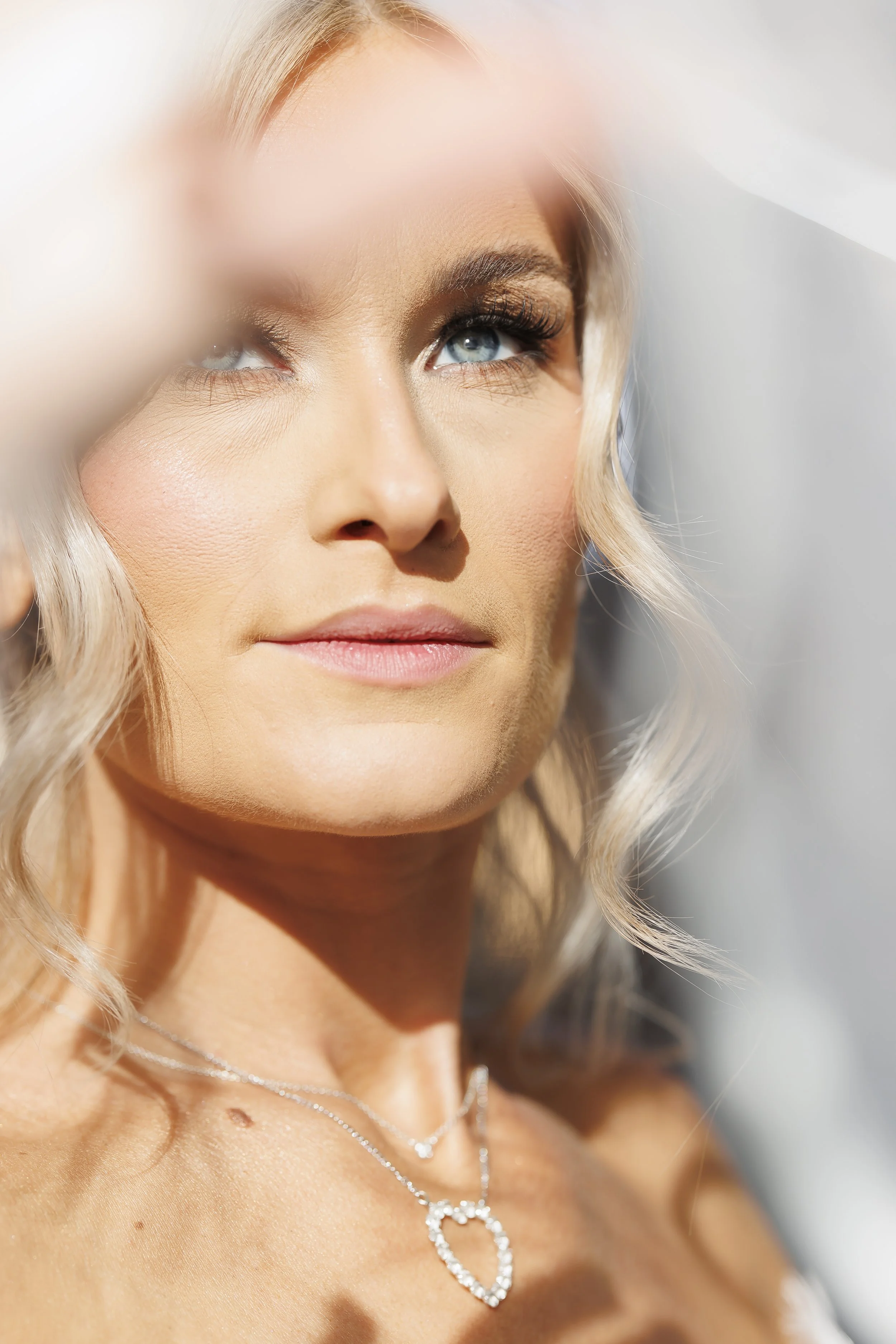 A woman with blonde hair, blue eyes, wearing makeup, and a silver necklace with a heart-shaped pendant, appears to be gazing upward in sunlight.