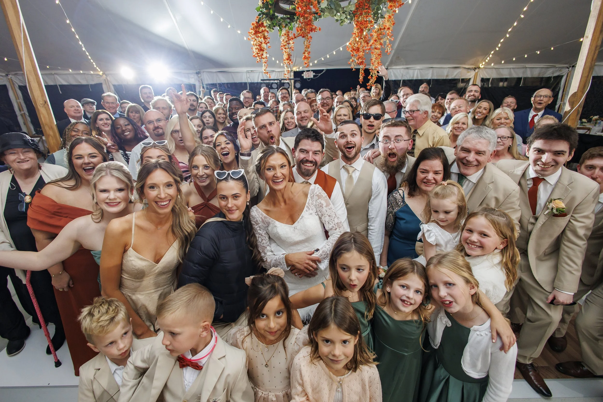 A large group of people at a wedding celebration, smiling and posing for a group photo inside a decorated tent with string lights and floral arrangements.