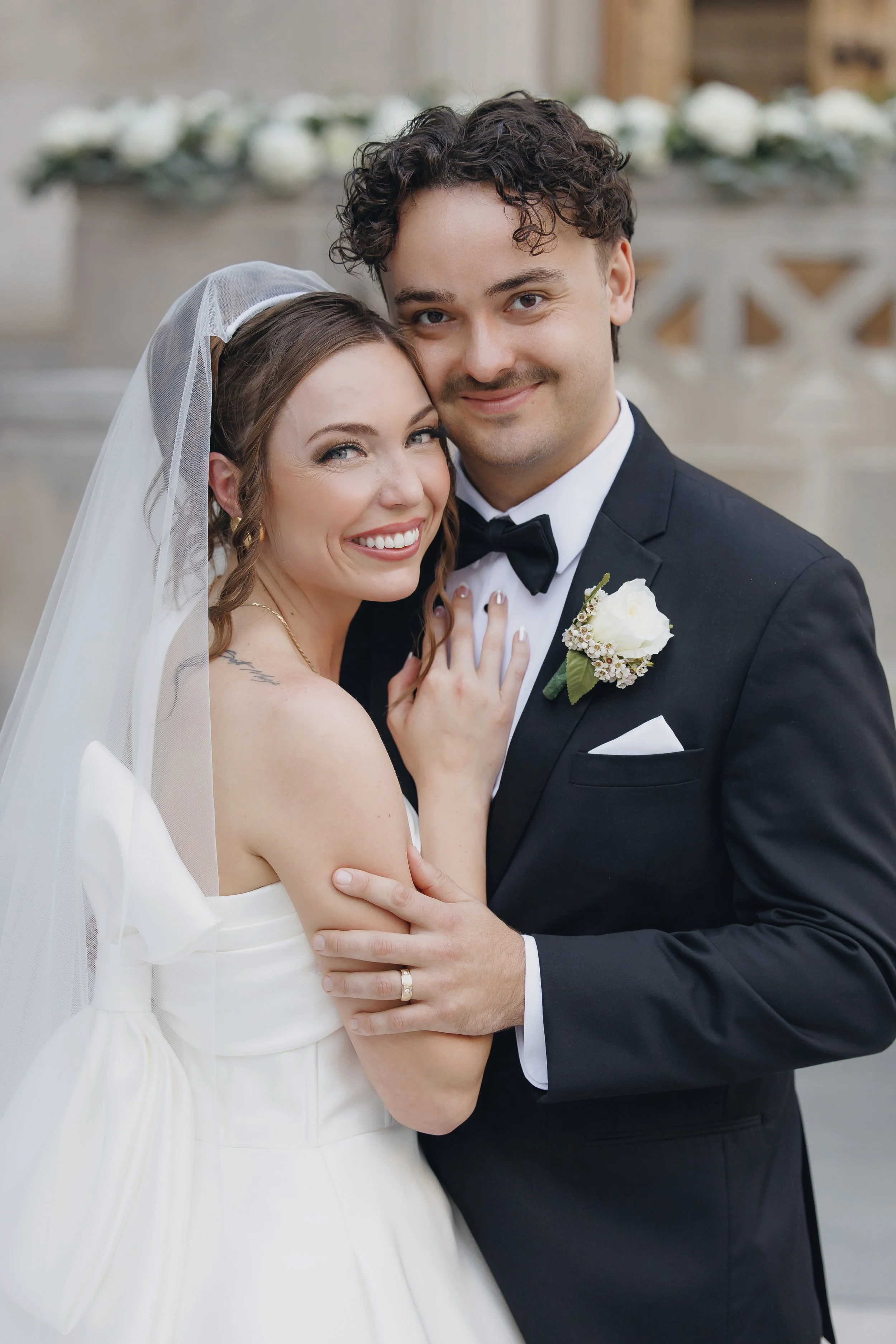 A bride and groom smiling and embracing on their wedding day, with the bride wearing a white dress and veil and the groom in a black tuxedo with a boutonniere.