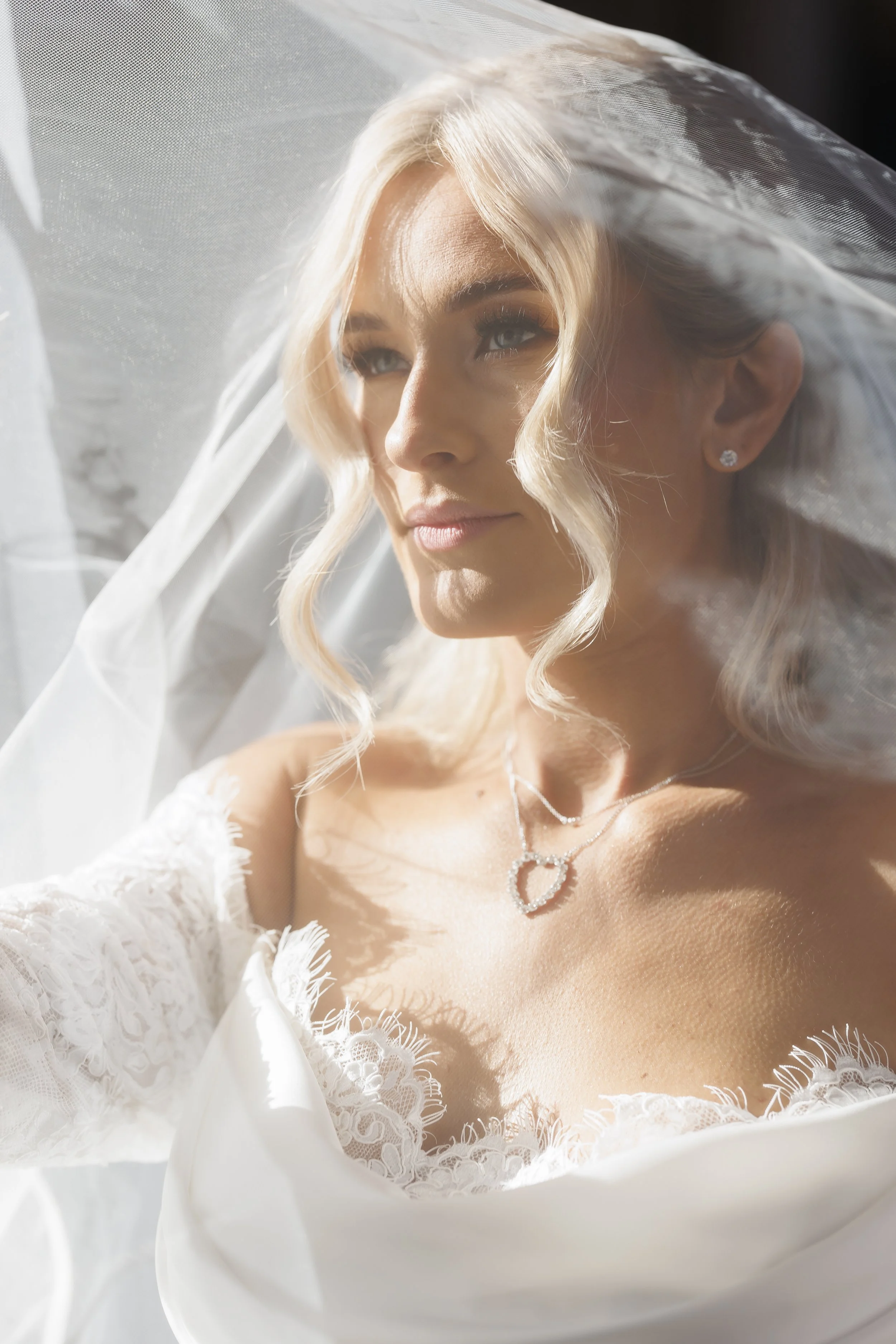 A bride with blonde hair in loose curls, wearing a white wedding dress with lace details, a necklace, and earrings, looking through bridal veil with soft sunlight