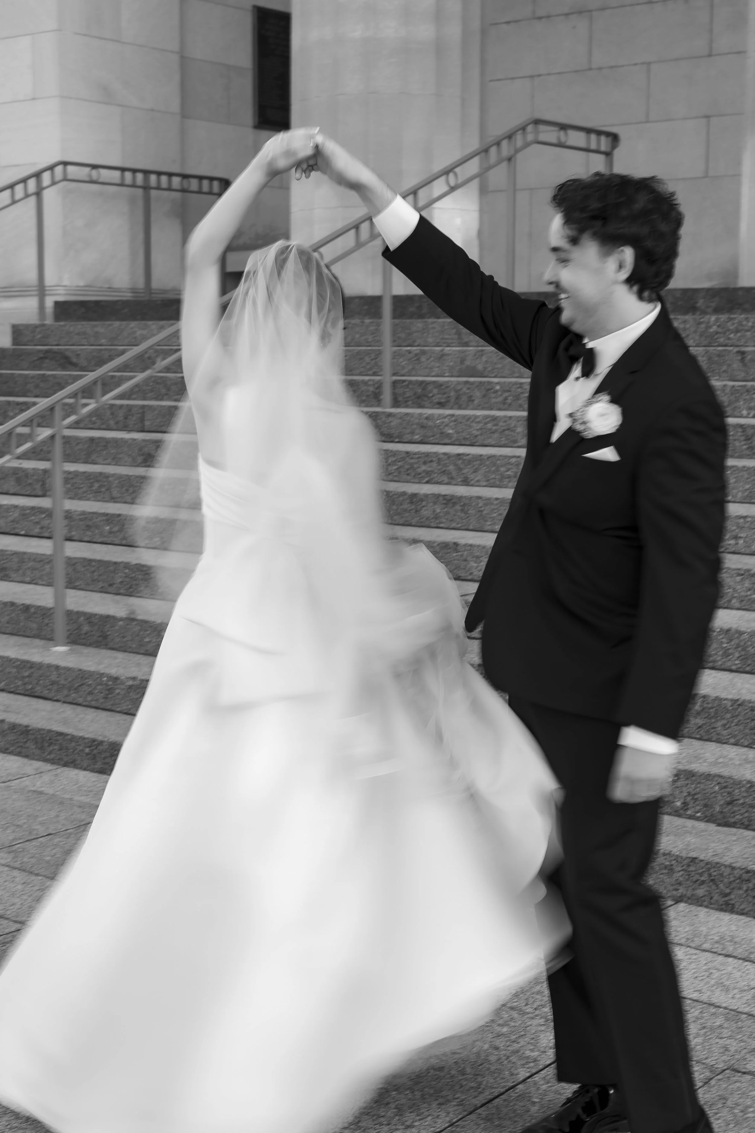 A bride and groom dancing outside on steps, with the bride's face obscured by her veil, black and white photo.