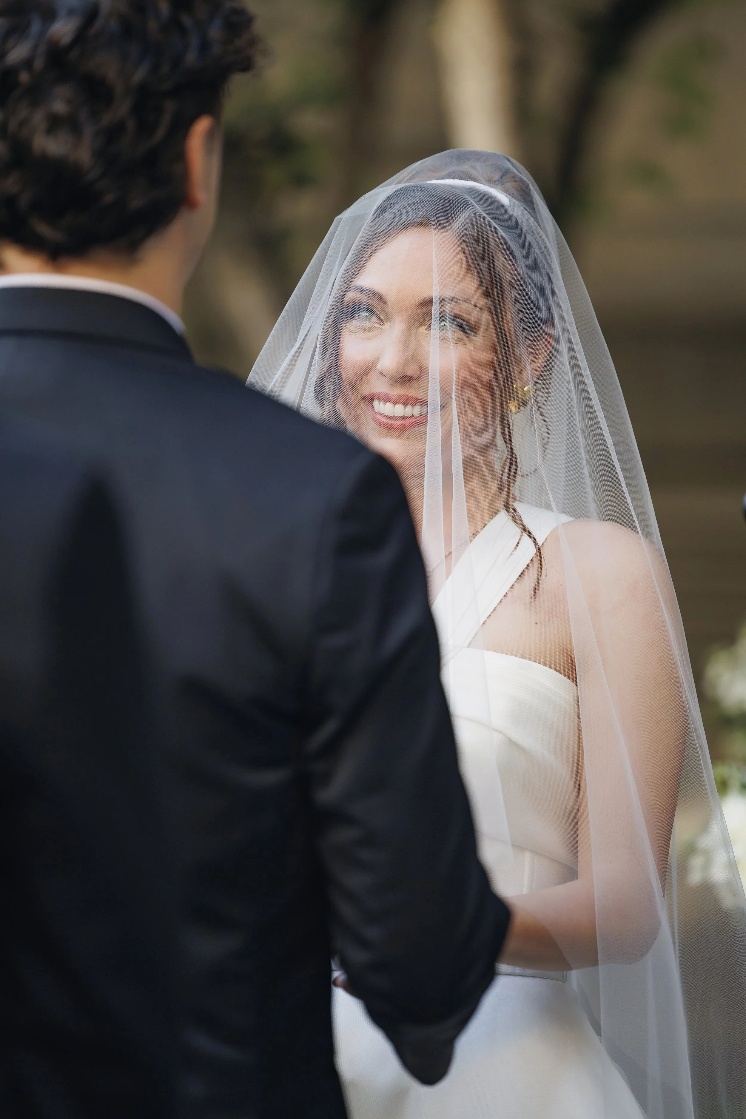 A bride and groom exchange vows outdoors, with the bride smiling at the groom through her sheer veil, surrounded by a blurred natural background.