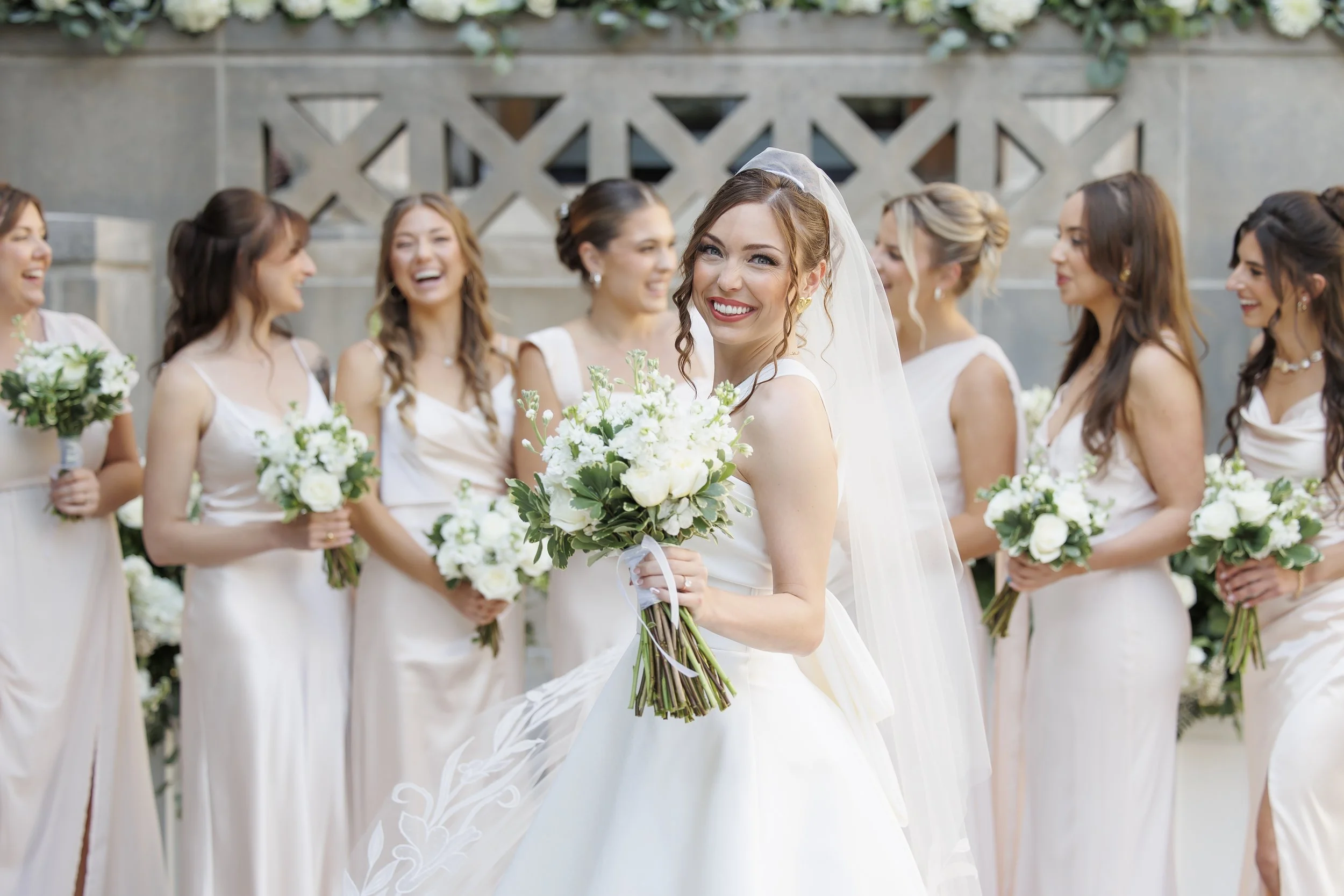 A bride with a veil holding a bouquet of white flowers, surrounded by bridesmaids in light-colored dresses, all smiling and standing outdoors in front of a decorative stone wall with greenery.