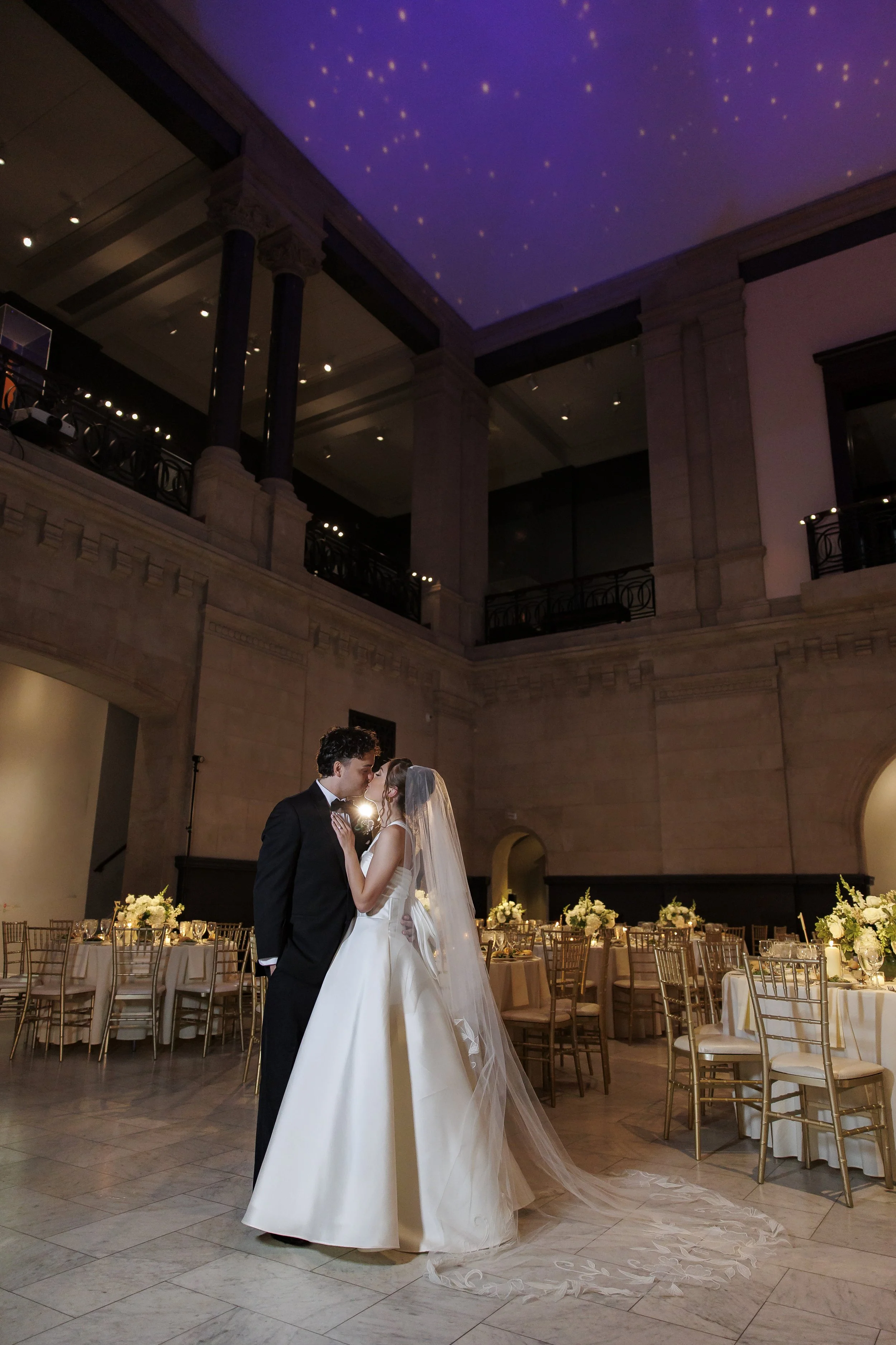 A bride and groom sharing a kiss in a wedding reception hall with decorated tables and a starry ceiling.