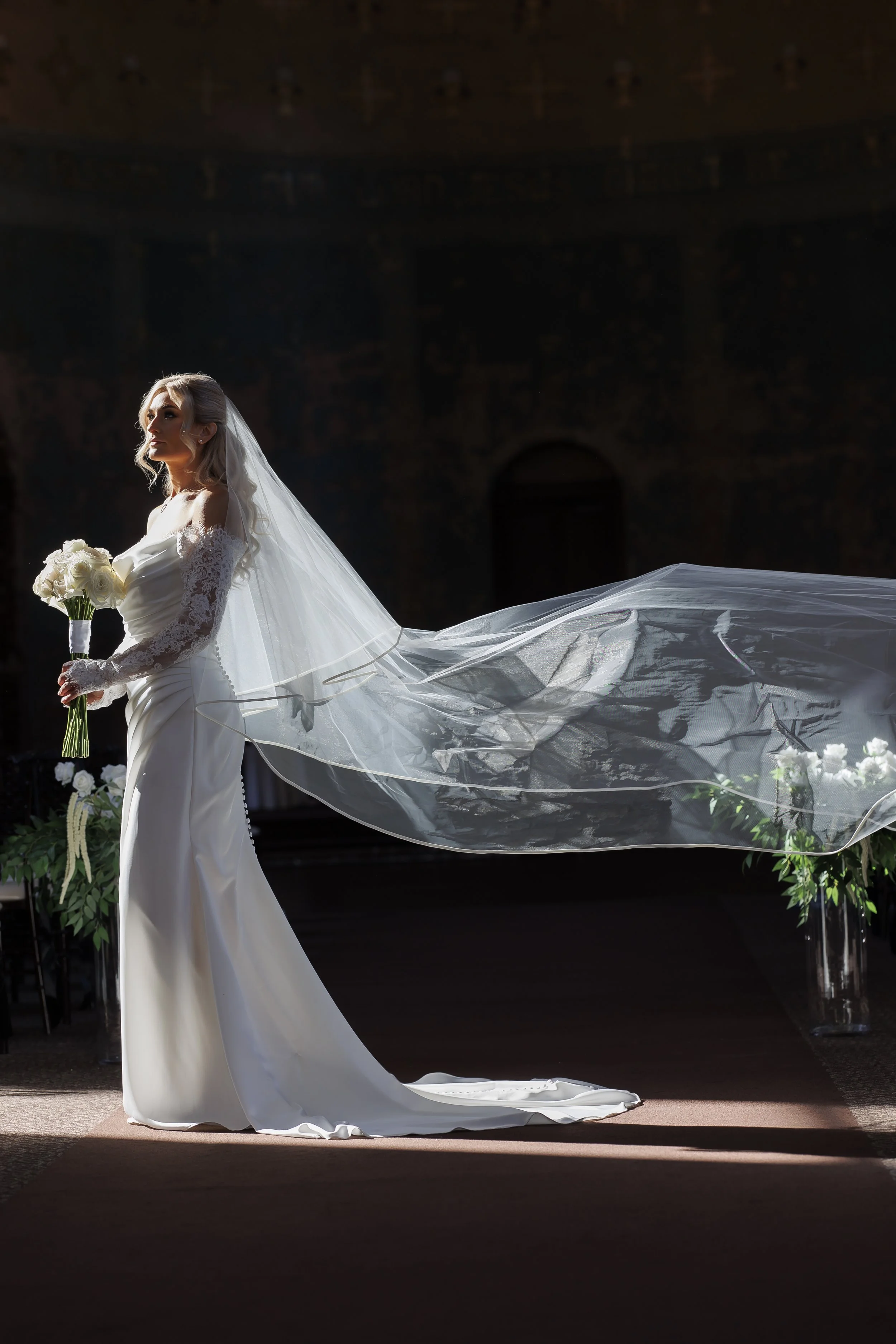A bride standing indoors holds a bouquet of white roses, dressed in an off-shoulder white wedding gown with lace sleeves, and wearing a veil flowing behind her during a wedding ceremony.