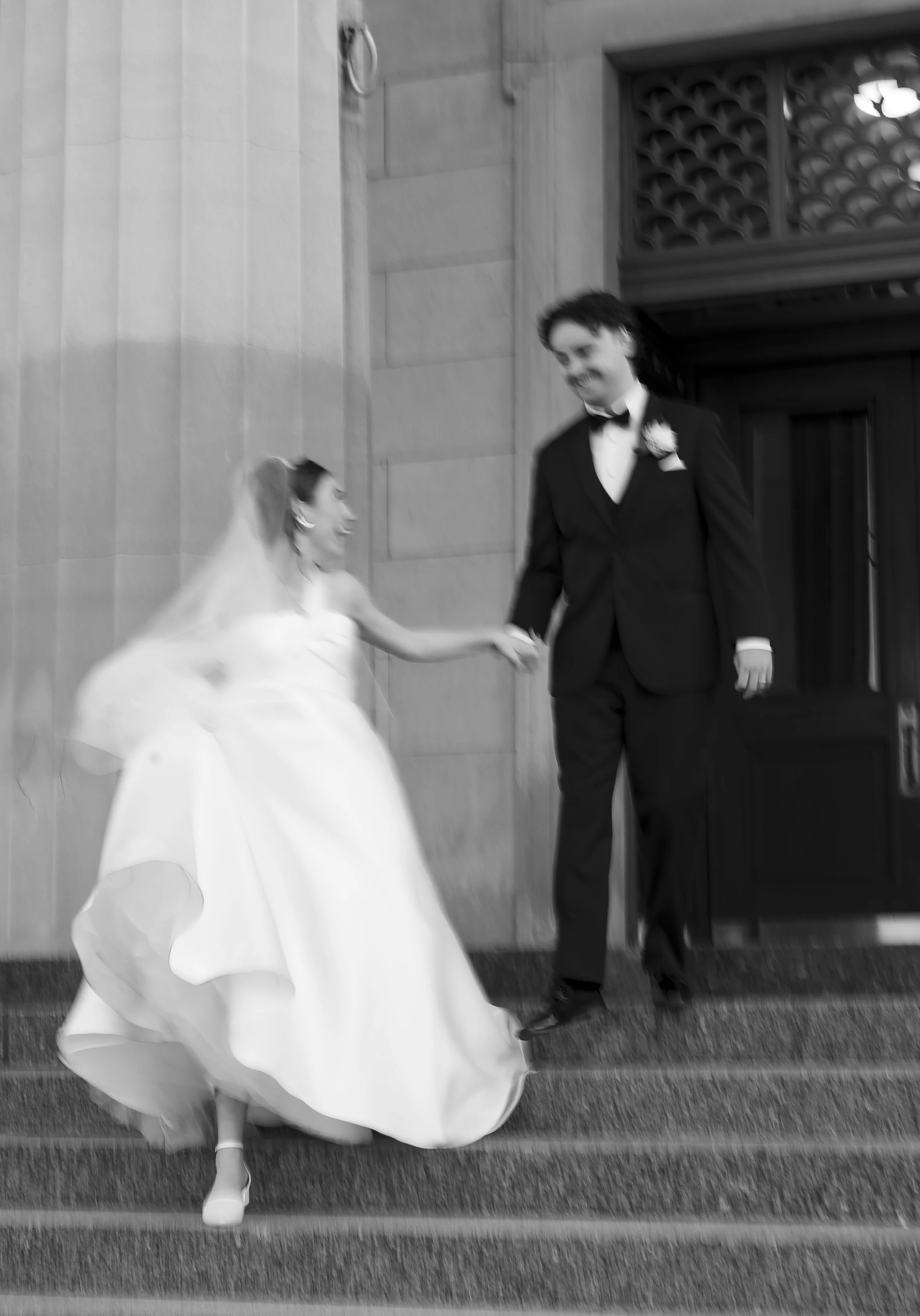 A bride and groom holding hands and smiling on the steps outside a church. The bride is wearing a wedding dress and veil, and the groom is dressed in a tuxedo.