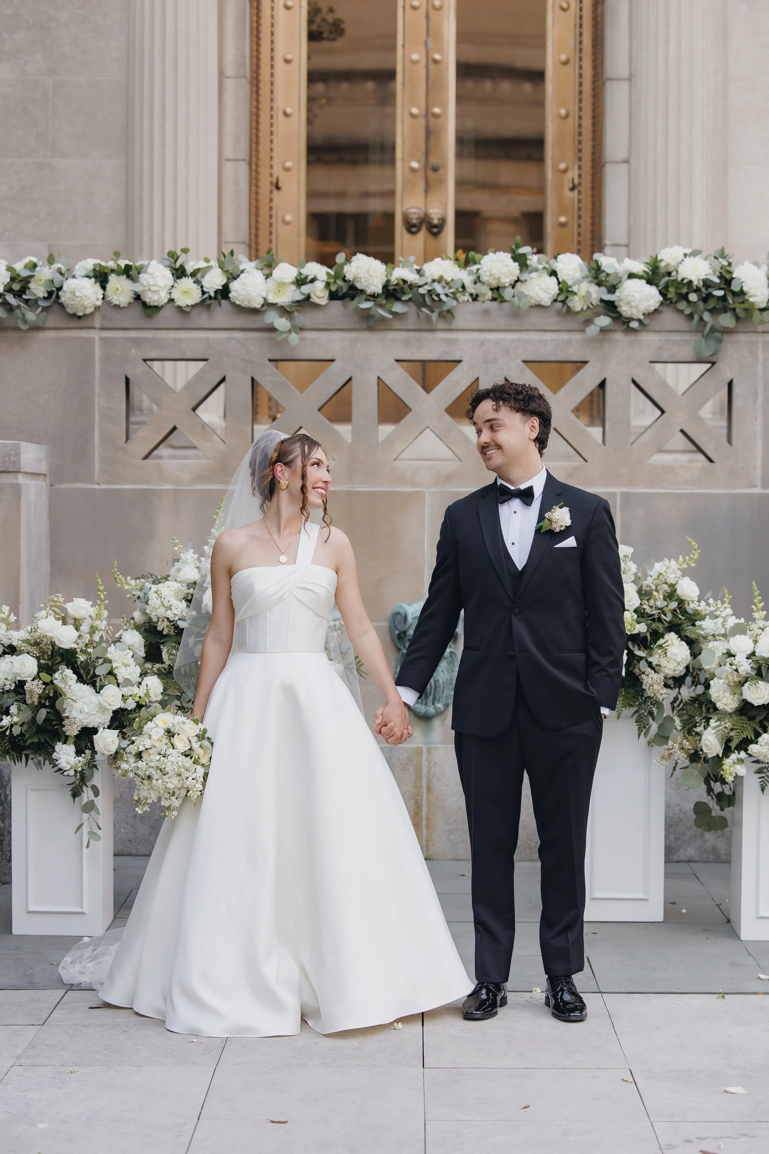 Bride and groom holding hands at their wedding ceremony, standing in front of floral arrangements and a stone wall with architectural details.