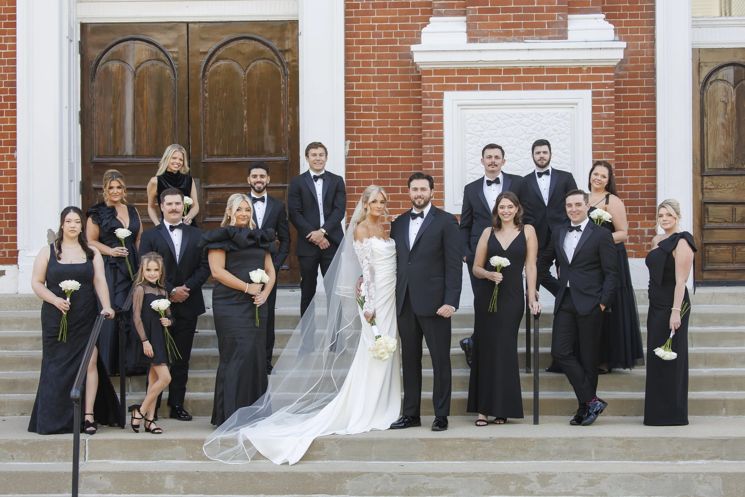 Wedding party on church steps with bride in white gown and groom in tuxedo surrounded by bridesmaids and groomsmen in black formal attire holding white roses.