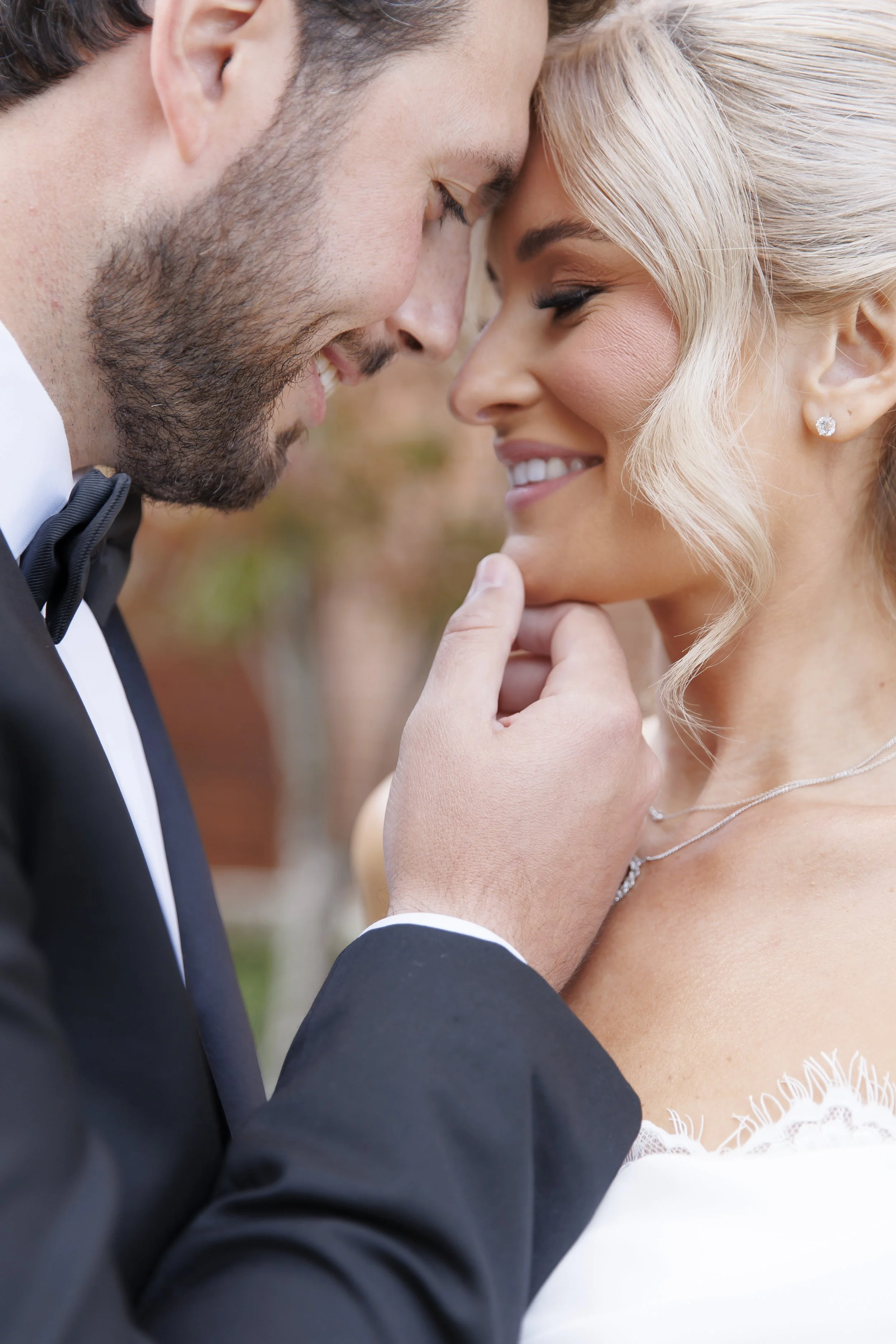 A newlywed couple in wedding attire sharing an intimate moment, with their foreheads touching and the groom gently holding the bride's chin, smiling softly.