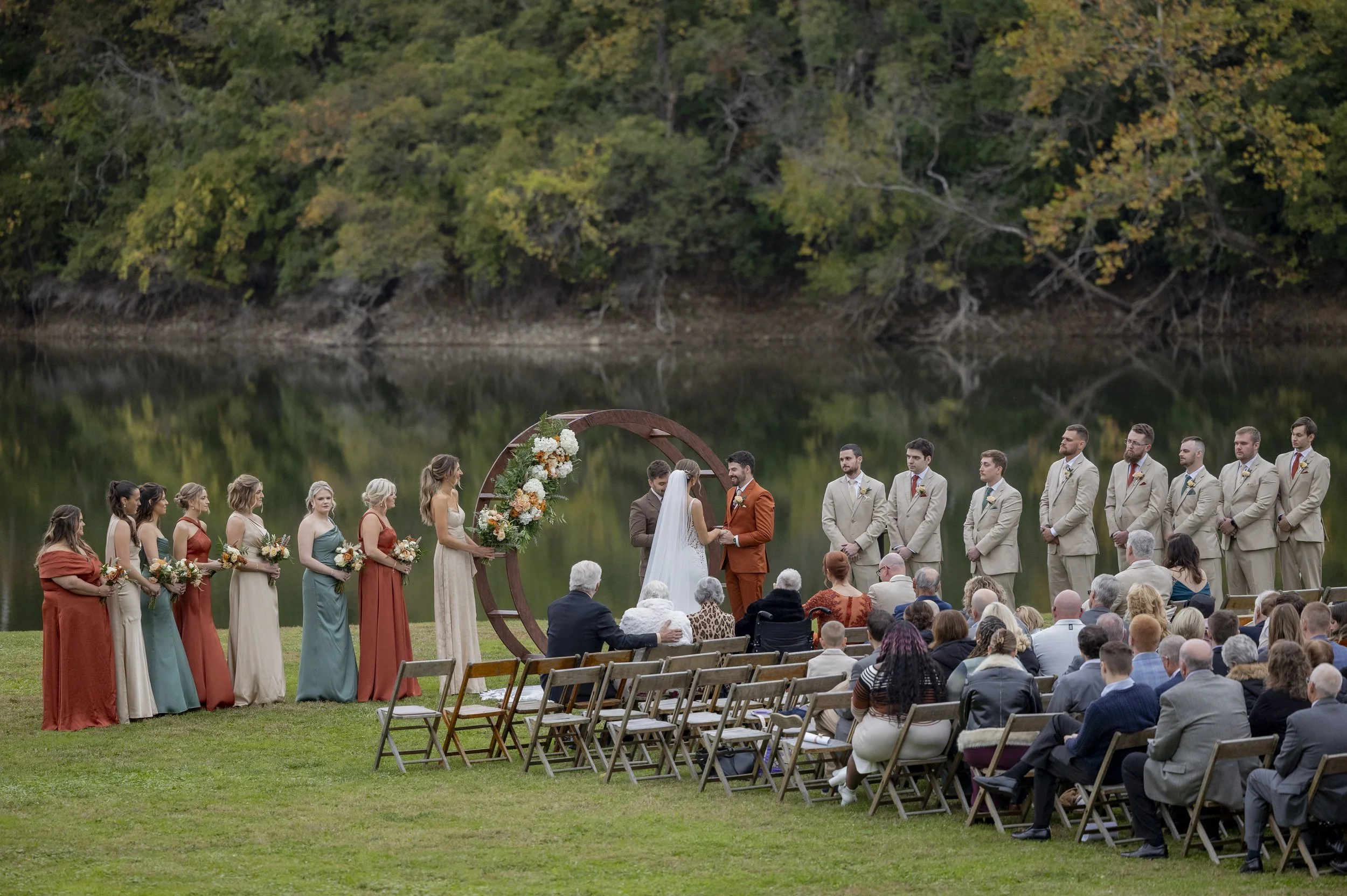 A wedding ceremony outdoors by a lake with a large group of guests seated on chairs. The couple is standing in front of a circular floral arch, exchanging vows. The bridal party is standing to the left and right of the couple, holding bouquets. The s
