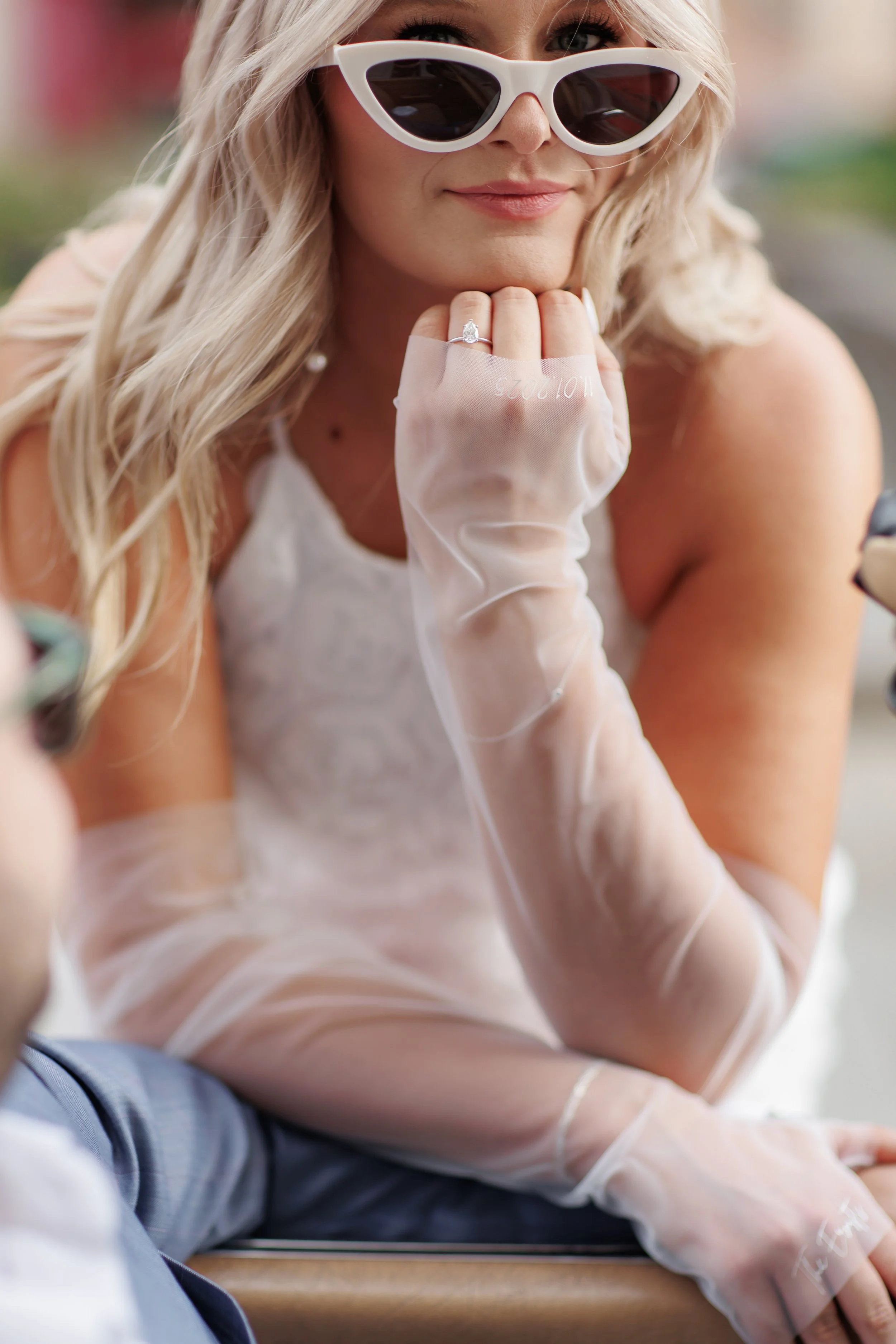 Close-up of a woman with long blonde hair wearing white cat-eye sunglasses and sheer gloves, resting her chin on her hand with a ring on her finger.