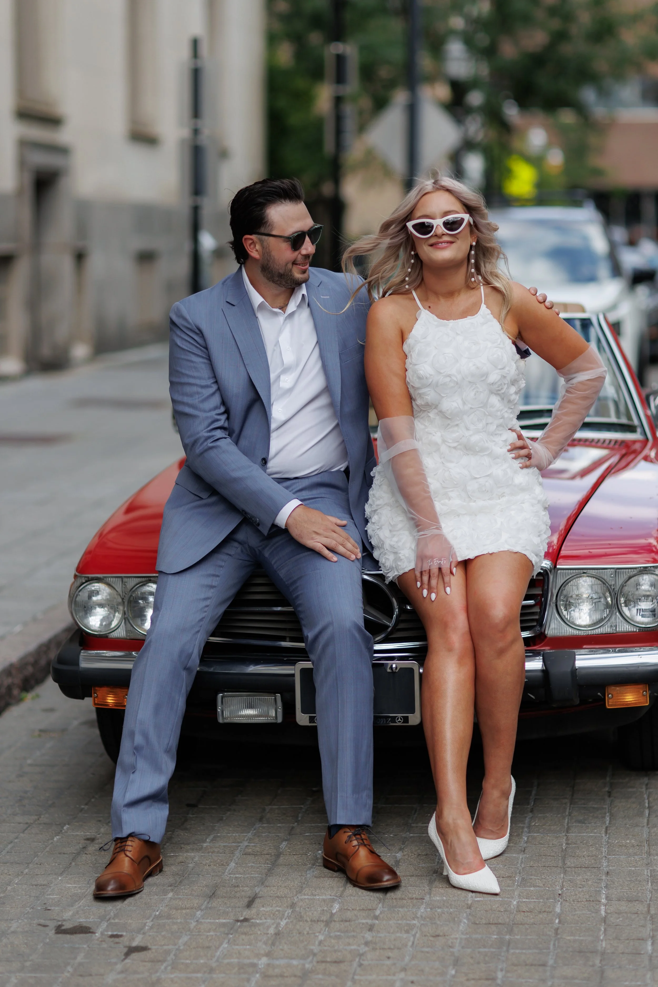 A fashionable couple sitting on the hood of a red vintage Mercedes-Benz car on a city street, smiling and enjoying each other's company.