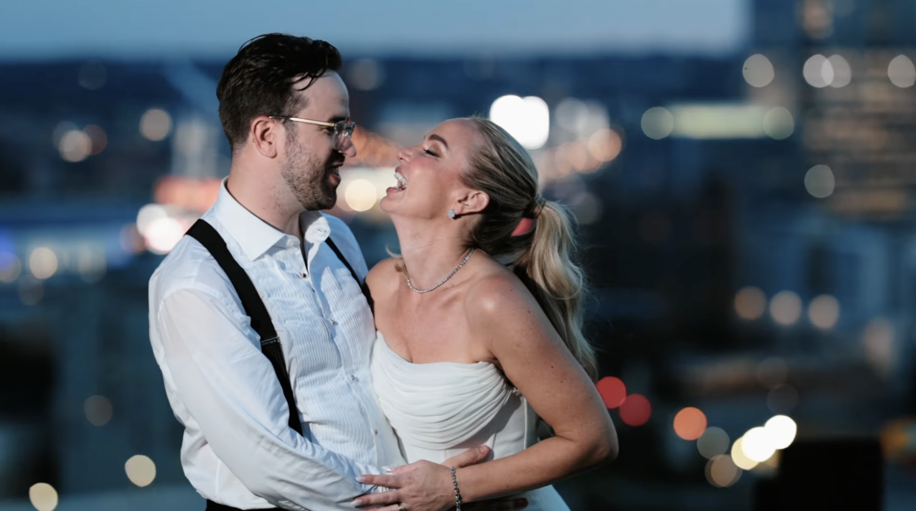 A couple embracing and smiling at each other with a city skyline and blurred lights in the background at dusk or night.